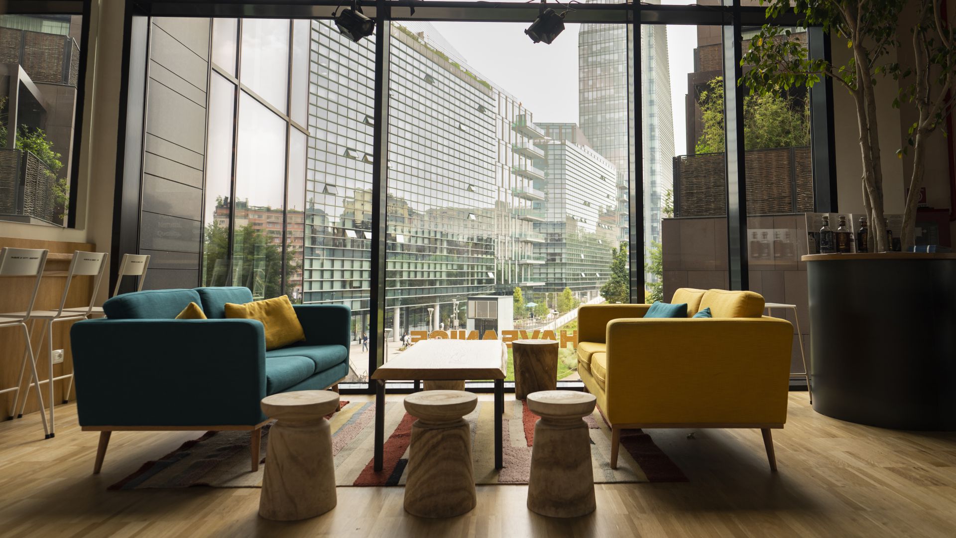 Moderna area lounge con divani blu e gialli, sgabelli in legno e tavolino da caffè su un tappeto colorato. Le grandi finestre a tutta altezza mostrano il paesaggio urbano, mentre i pezzi della Tripstillery Art Collection aggiungono un tocco creativo.