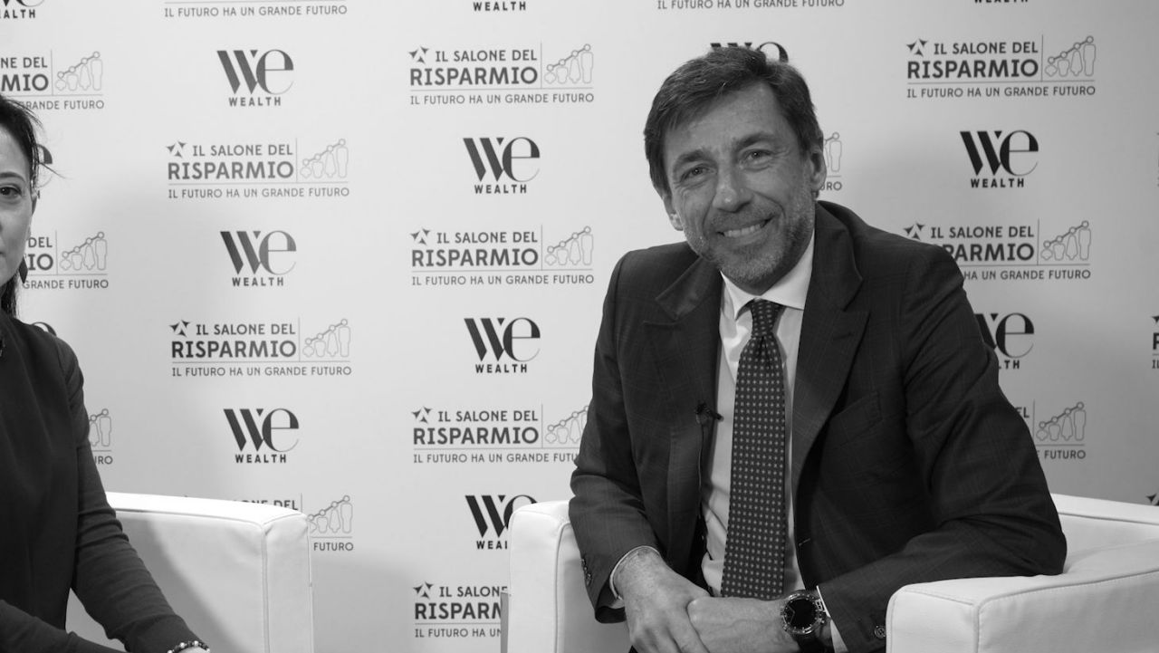 Un uomo in giacca e cravatta siede e sorride durante un'intervista a un evento con i loghi "We Wealth" e "Il Salone del Risparmio" sullo sfondo. Il braccio di un'altra persona è parzialmente visibile sulla sinistra. L'immagine è in bianco e nero.
