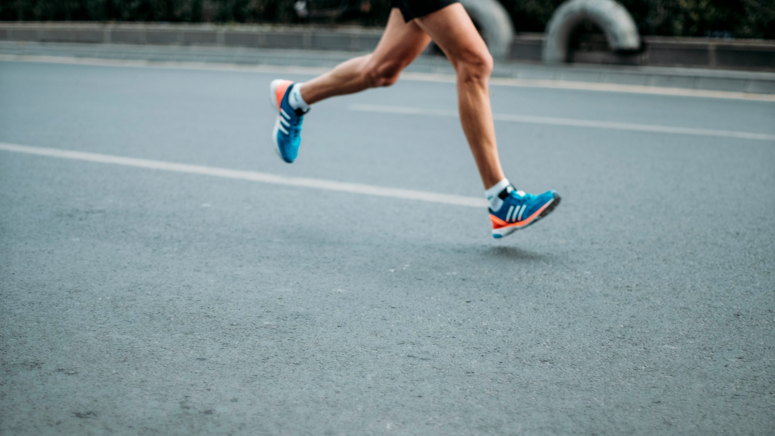 Una persona che indossa scarpe sportive blu e pantaloncini neri sta correndo su una strada vuota, con le sole gambe visibili a metà corsa.