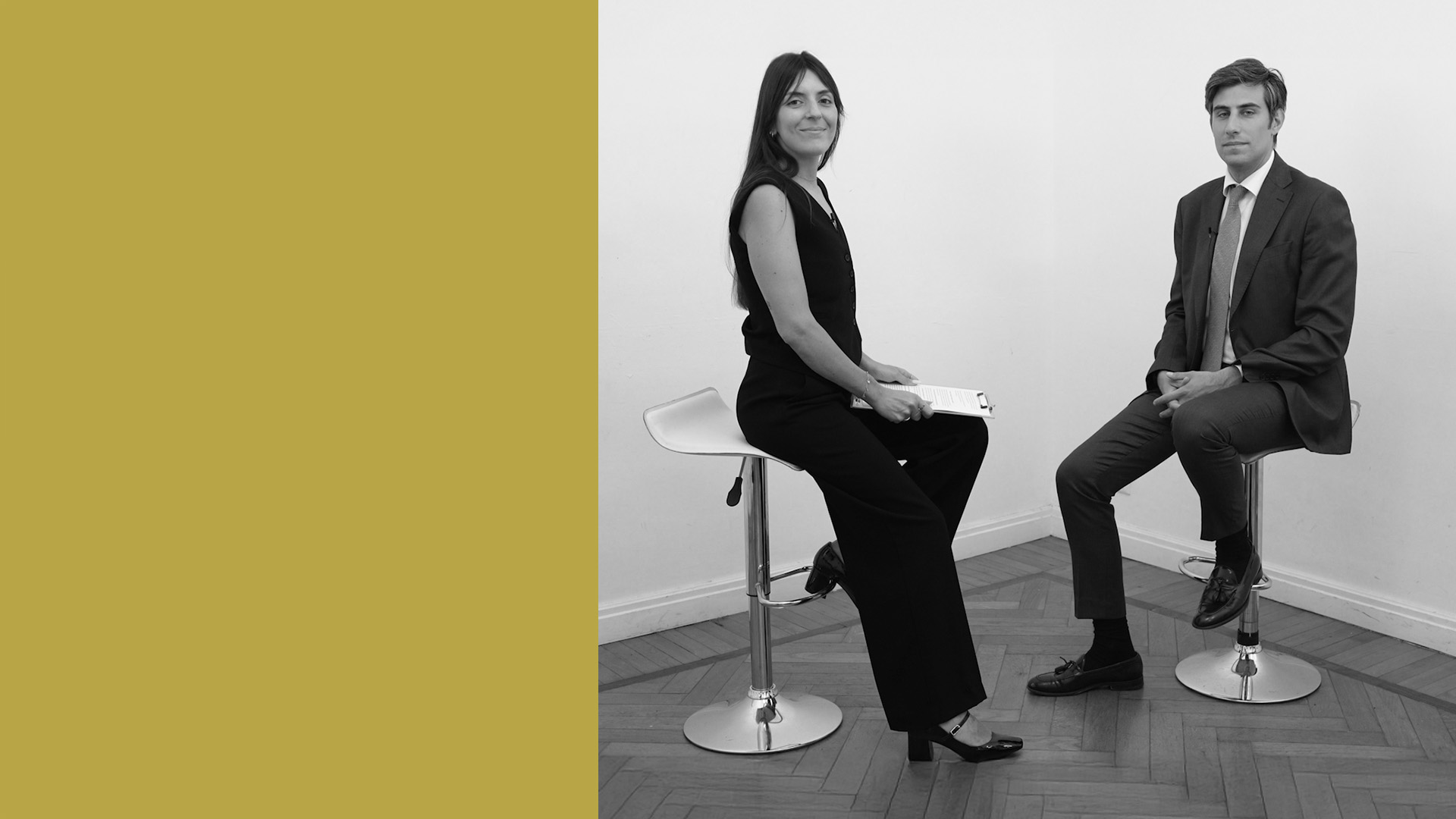 Una donna con dei documenti in mano e un uomo in giacca e cravatta sono seduti su degli sgabelli da bar, uno di fronte all'altro, in una stanza minimalista, con un grande blocco giallo sul lato sinistro dell'immagine.