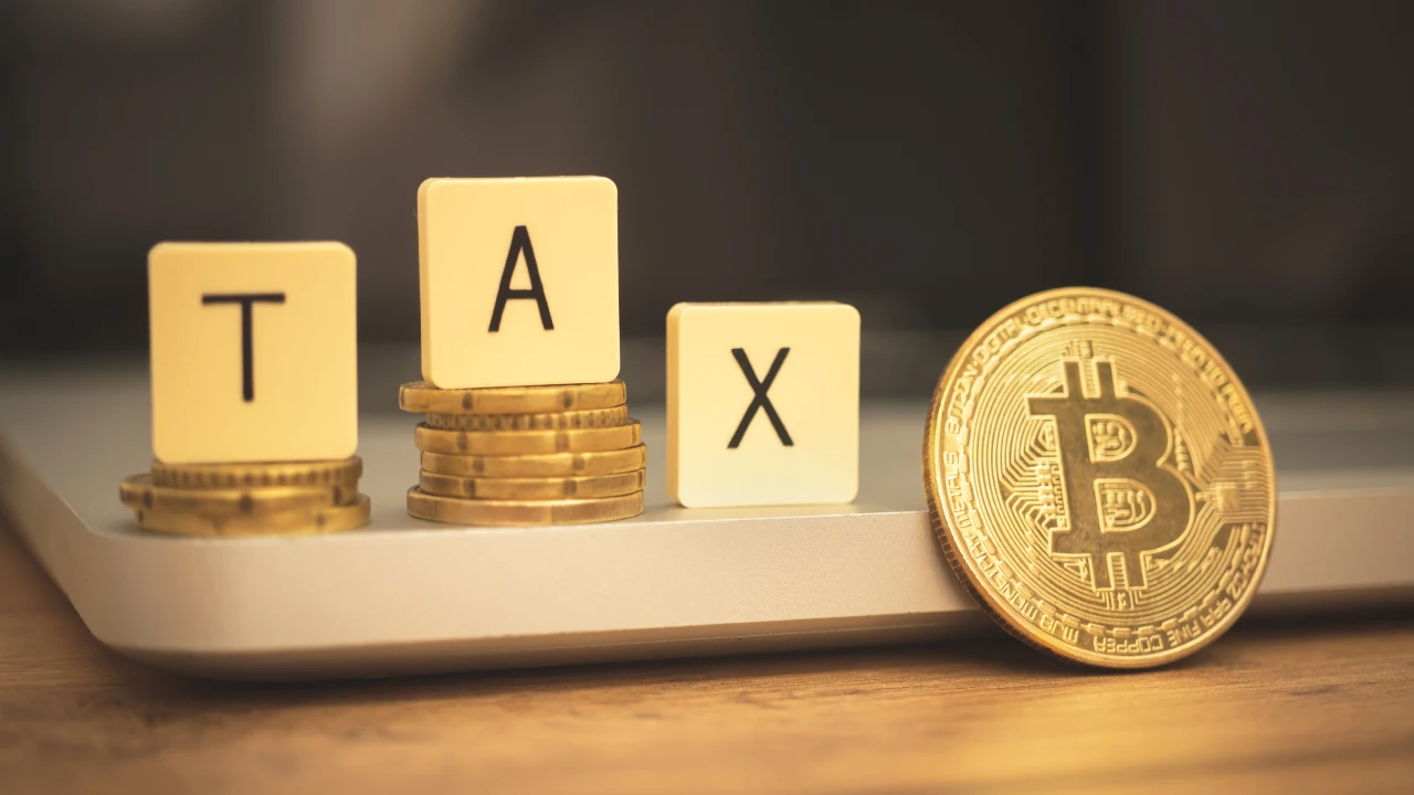 Tre pile di monete con sopra dei blocchi di lettere che compongono la parola "TASSA", accanto a grande moneta Bitcoin d'oro, il tutto posizionato sulla superficie di un computer portatile, a simboleggiare l'intersezione tra criptovalute e tassazione.