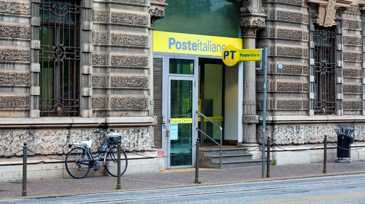 Ingresso di un ufficio di Poste Italiane con cartello giallo su un edificio in pietra decorata, una bicicletta parcheggiata nelle vicinanze ed elementi stradali come dissuasori e un cestino dei rifiuti di fronte.