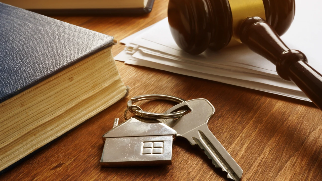 Un portachiavi a forma di casa con chiavi è appoggiato su una scrivania di legno accanto a un libro di legge, a documenti legali e al martelletto di un giudice, a simboleggiare il diritto immobiliare o le questioni legali che coinvolgono il coniuge.