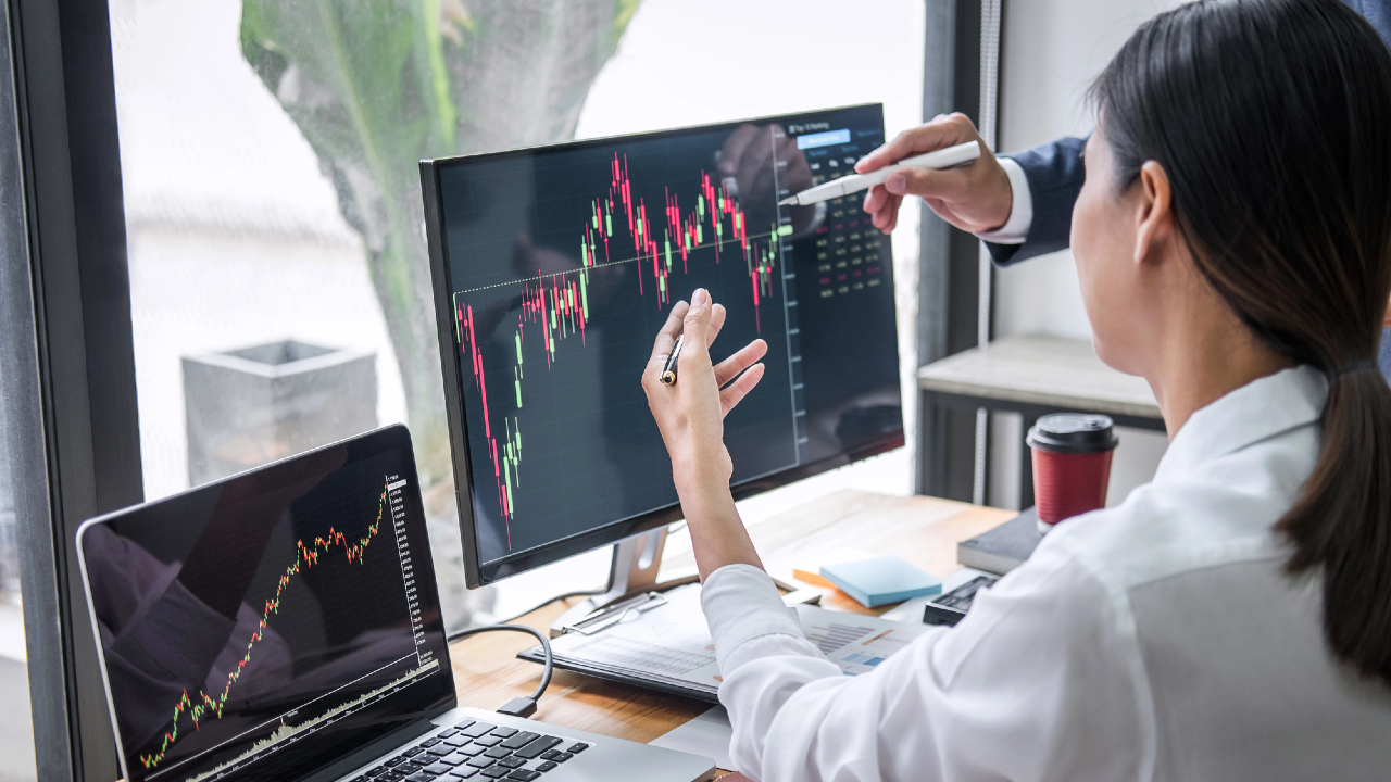 Due persone, sedute a una scrivania, analizzano grafici finanziari su un computer e un portatile; una indica lo schermo con una penna, discutendo delle performance degli ETF, delle tendenze del mercato azionario e dei dati.
