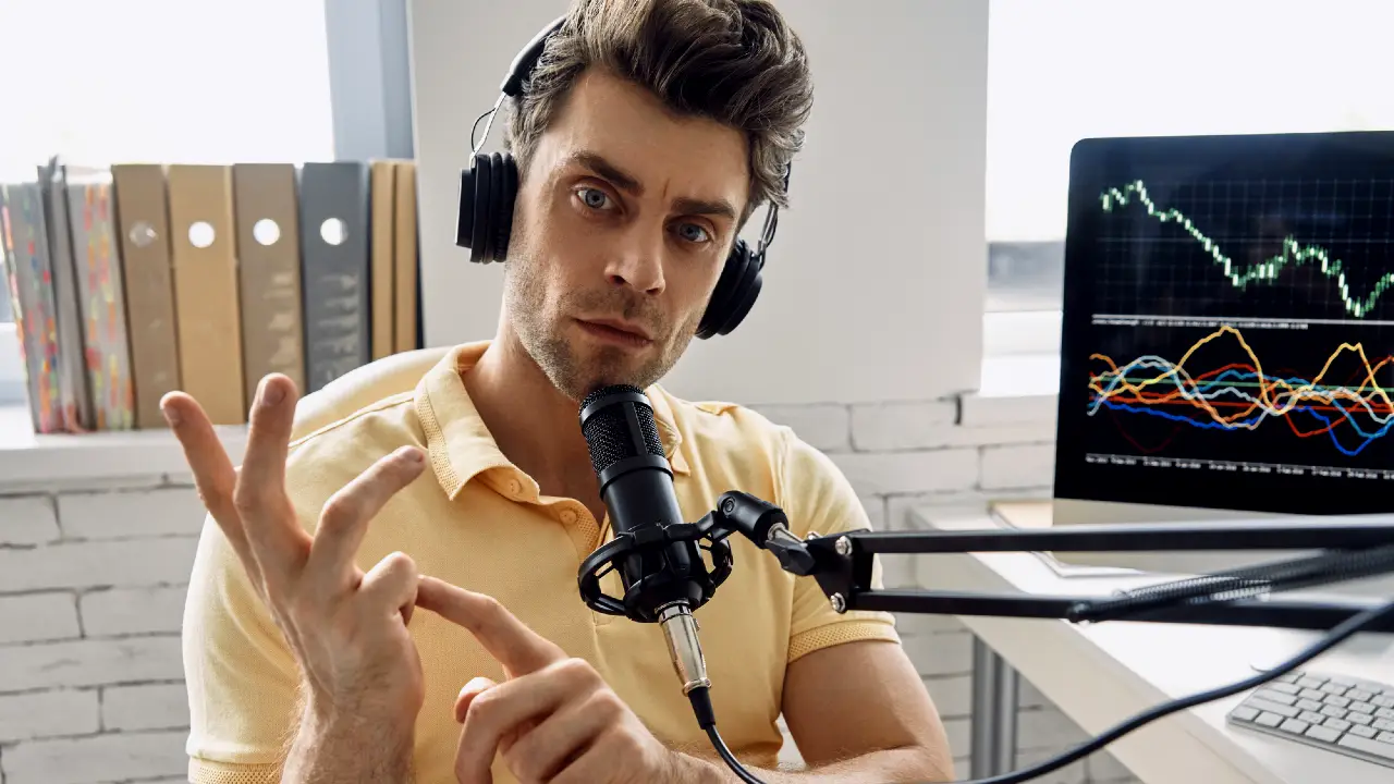 Un uomo con le cuffie e una polo gialla parla in un microfono, usando le dita per contare i punti. Dietro di lui, un monitor di computer mostra grafici a linee colorate. Sullo sfondo si vedono scaffali con cartelle.