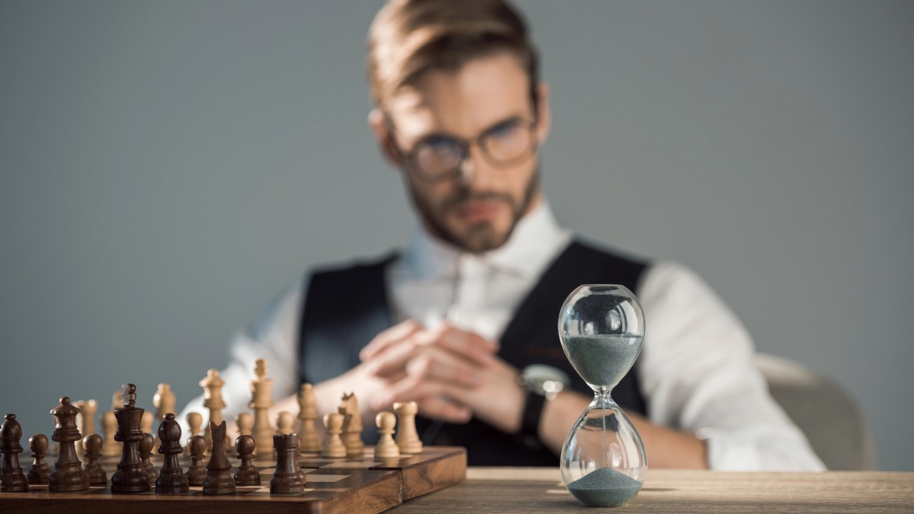 Un uomo con gli occhiali siede pensieroso a un tavolo, vicino a una scacchiera e a una clessidra, con sguardo pensieroso cercando di capire la mossa da fare, ossia la scelta di holding da privilegiare in tema di pianificazione strategica.