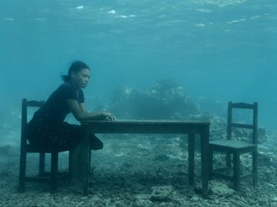 Una persona siede da sola a un tavolo di legno con due sedie sott'acqua, circondata da un fondale roccioso e dalla luce diffusa del sole che filtra attraverso l'acqua sovrastante, riflettendosi su bnp volontariato sotto le onde tranquille.