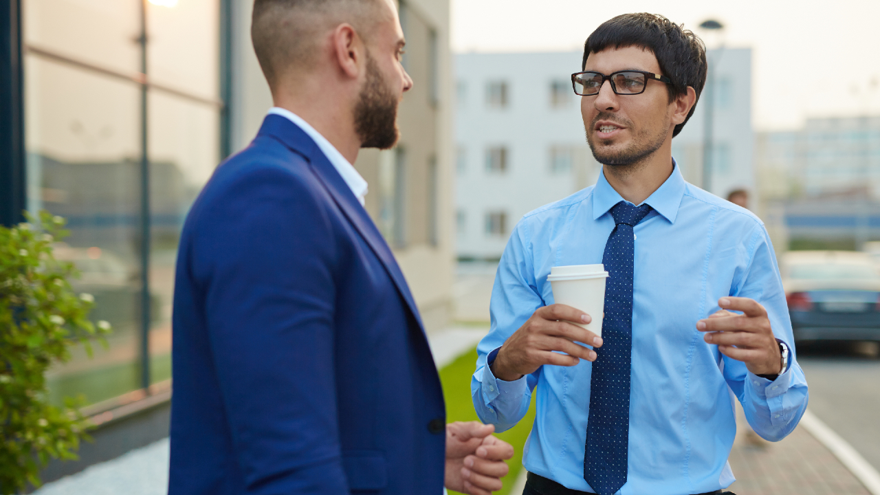 Due uomini in abiti da lavoro, probabilmente banchieri, sono all'esterno e stanno conversando. Uno tiene in mano una tazza di caffè e gesticola animatamente, mentre l'altro ascolta con attenzione.
