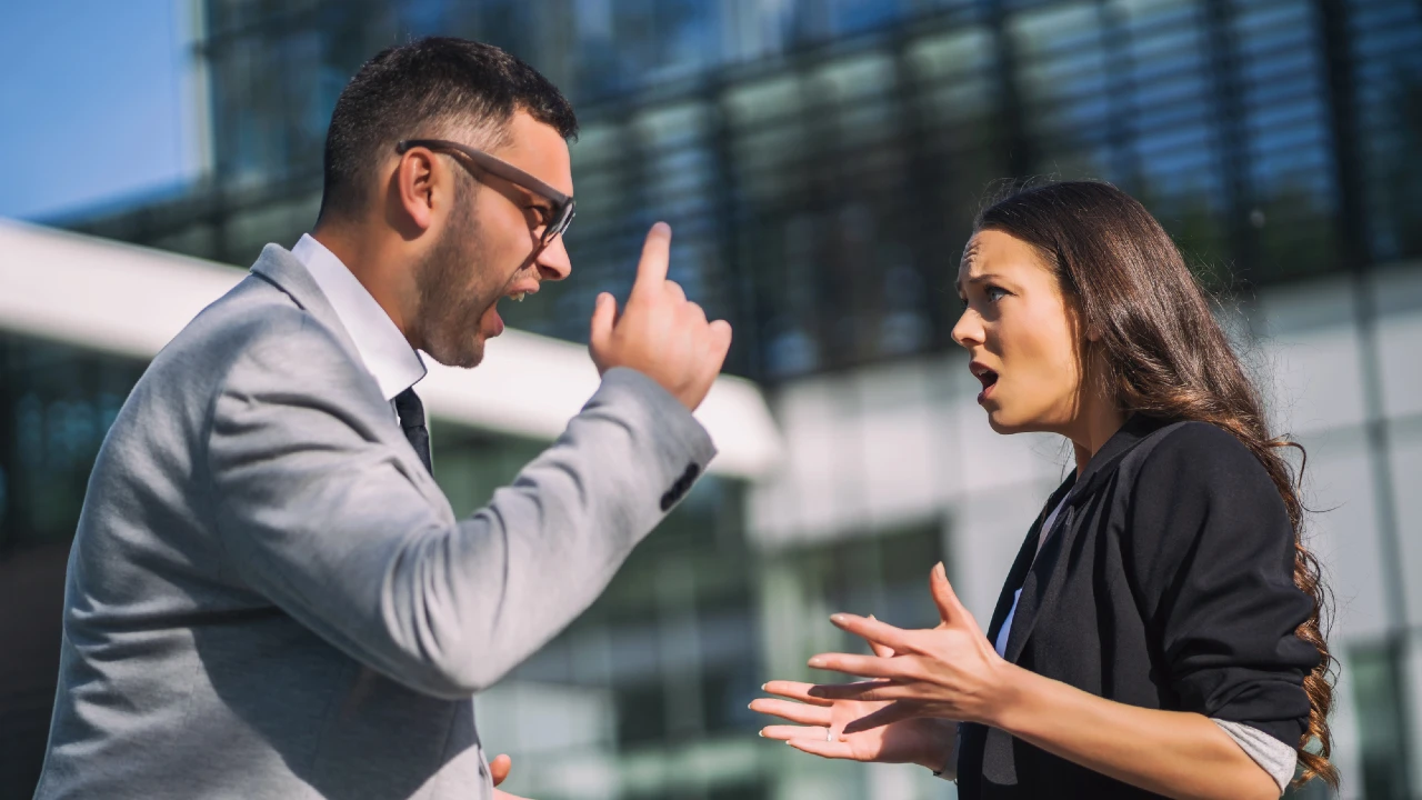 Un uomo e una donna in abiti da lavoro stanno avendo un'accesa discussione all'aperto, probabilmente legata a questioni di lavoro e mobbing, gesticolando con le mani e apparendo turbati. Sullo sfondo, un moderno edificio in vetro.