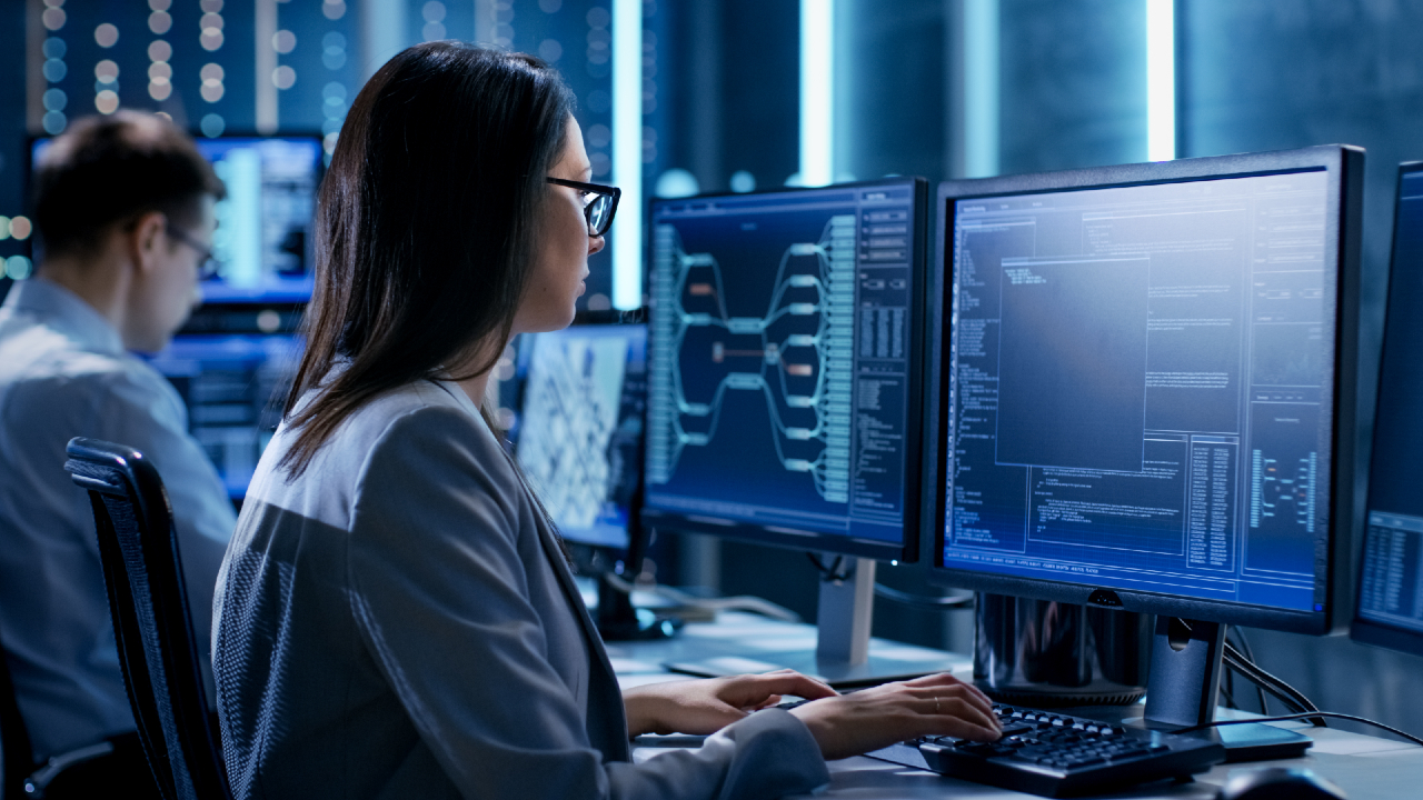 Una donna con gli occhiali analizza i dati di Azioni a una scrivania con diversi monitor che visualizzano codice in un ufficio moderno e tecnologico. Un'altra persona lavora a una scrivania separata sullo sfondo.