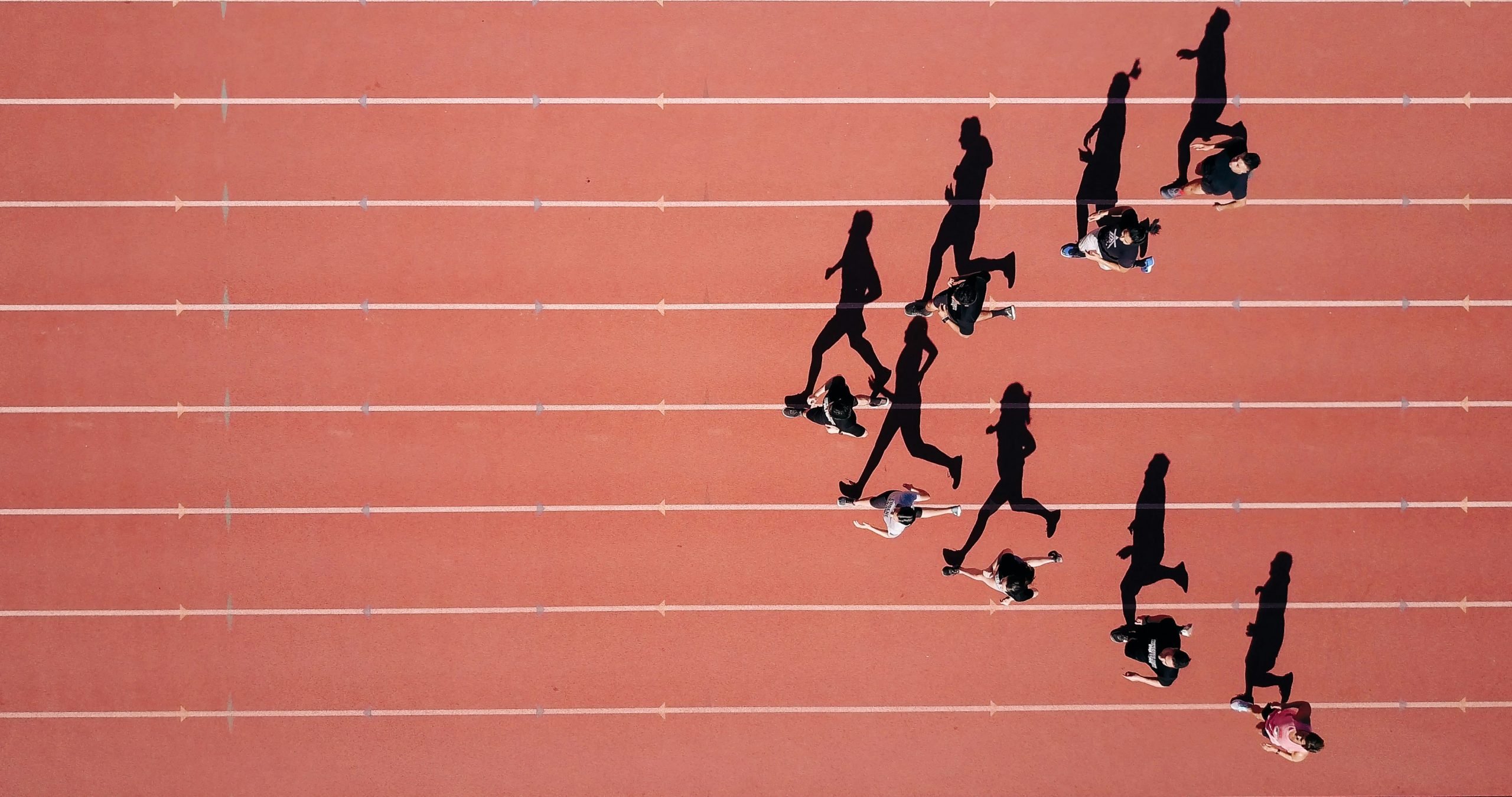 Vista aerea di un gruppo di persone che corrono su una pista di atletica rossa, proiettando lunghe ombre sulle corsie alla luce del sole.