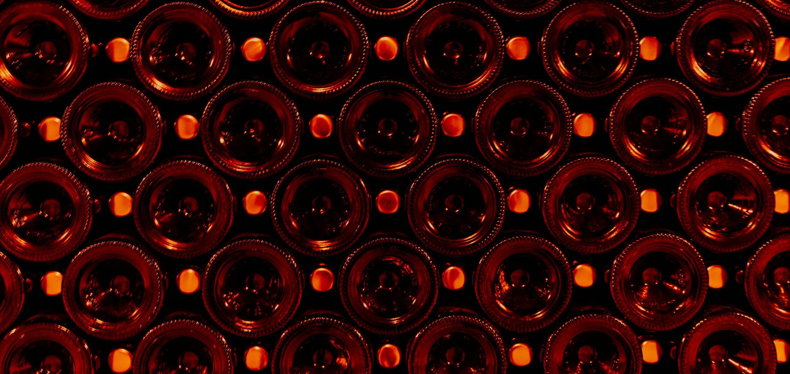 Sono raffigurate file di bottiglie di vino scuro impilate orizzontalmente con la base rivolta verso l'esterno, illuminate da una calda luce rosso-arancio che mette in risalto i motivi circolari e i tappi delle bottiglie.