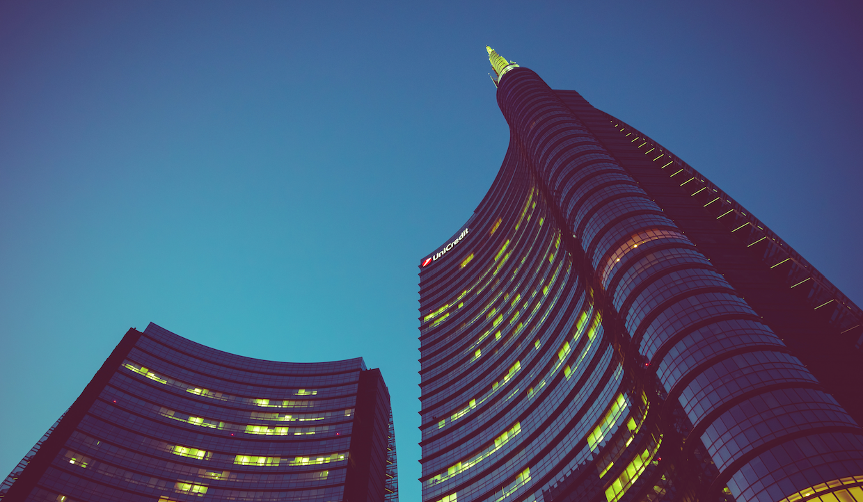 Due moderni grattacieli di vetro con finestre illuminate si vedono dal basso contro un cielo terso al tramonto. Un edificio è più alto e ha una guglia con la punta luminosa. L'atmosfera è tranquilla e urbana.