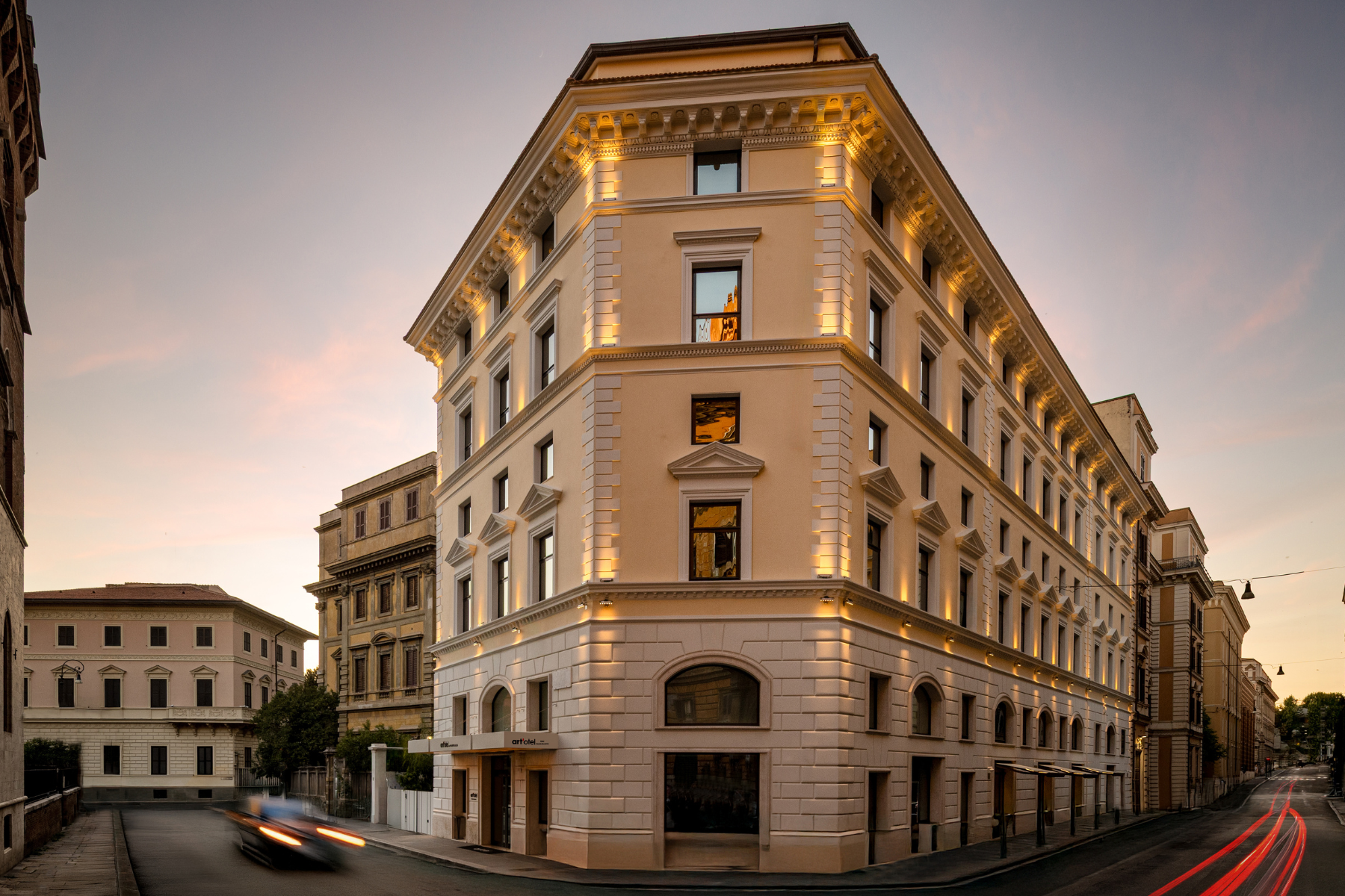 Un elegante edificio d'angolo a più piani con una calda illuminazione esterna, ampie finestre e dettagli architettonici classici, situato in una strada tranquilla al tramonto con le luci sfocate delle auto che passano.