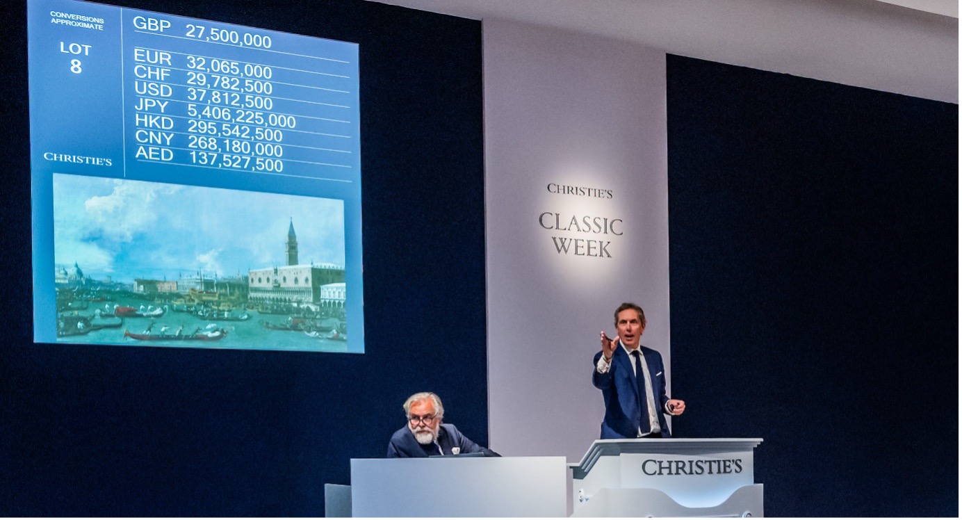 Un banditore d'asta gesticola mentre conduce una vendita alla Settimana Classica di Christie's, con conversioni di valuta e un dipinto da record di Canaletto visualizzato su un grande schermo alle sue spalle. Un'altra persona è seduta a una scrivania accanto al podio.