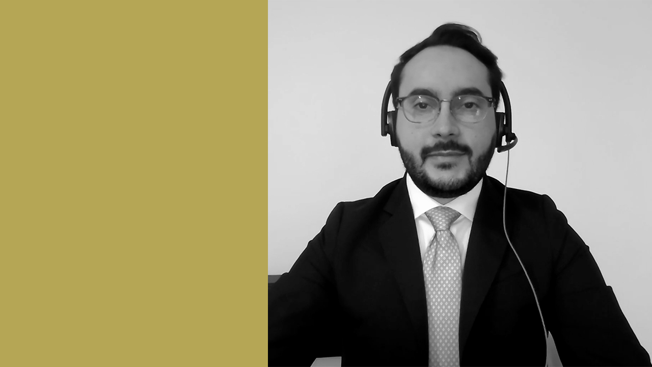 Un uomo che indossa occhiali, abito e cravatta è seduto di fronte alla telecamera con una cuffia in testa. Lo sfondo è semplice, con un grande blocco color oro sul lato sinistro e un'immagine in scala di grigi dell'uomo sulla destra.