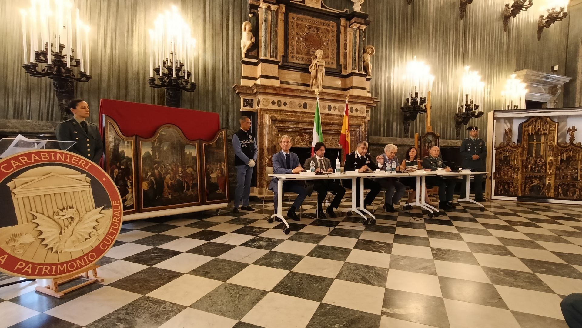 Un gruppo formale di sette persone siede a un lungo tavolo in una sala ornata con pavimenti a scacchi, circondata da opere d'arte e statue storiche. Ufficiali in uniforme sono in piedi nelle vicinanze e un grande cartello dei Carabinieri "Tutela Patrimonio Culturale" è esposto.