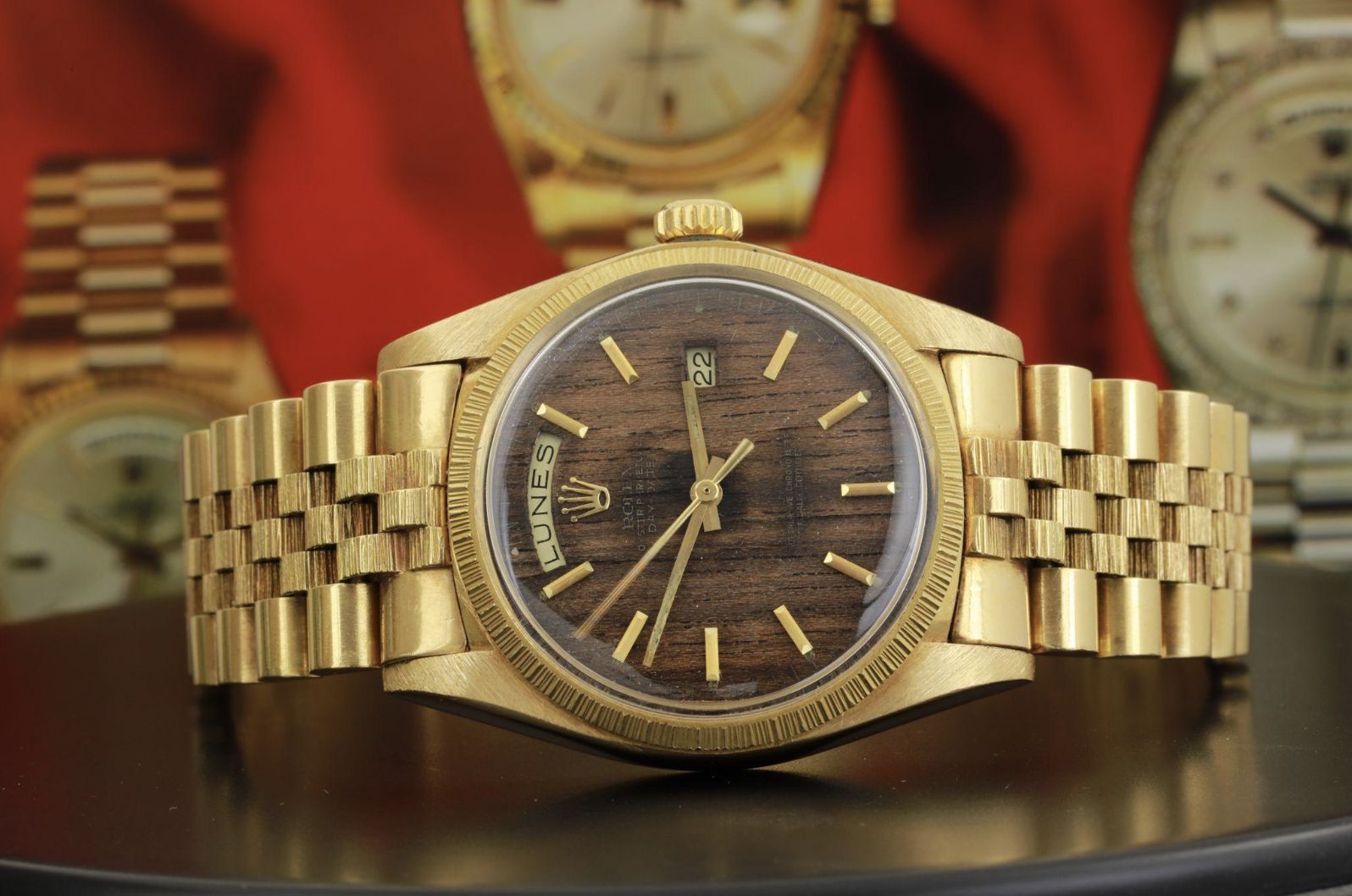 Orologio da polso in oro con quadrante marrone con motivo a legno, indici in oro e datario, con bracciale in oro abbinato. Sullo sfondo rosso sono visibili orologi di lusso sfocati.