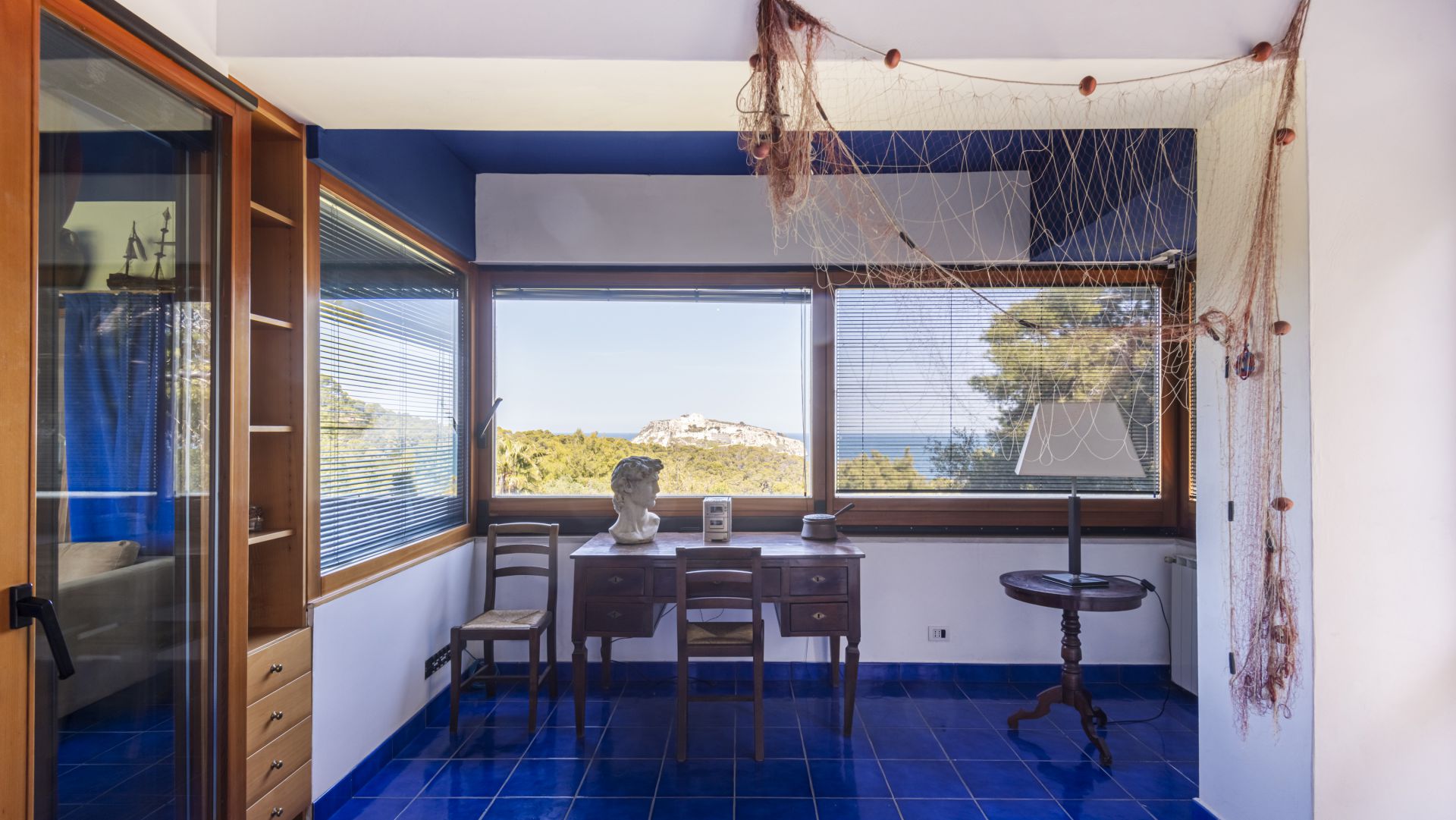 Una stanza luminosa con pavimento in piastrelle blu, scrivania e sedia in legno accanto a una grande finestra, un busto scultura sulla scrivania e reti da pesca appese al soffitto. All'esterno, una vista panoramica sugli alberi e su una collina lontana.