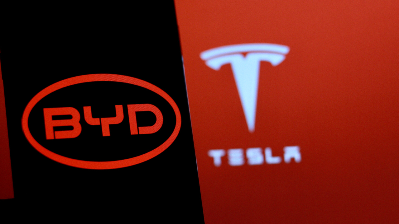 Primo piano dei loghi illuminati di BYD e Tesla su uno sfondo rosso e nero, che rappresentano i due produttori di veicoli elettrici.