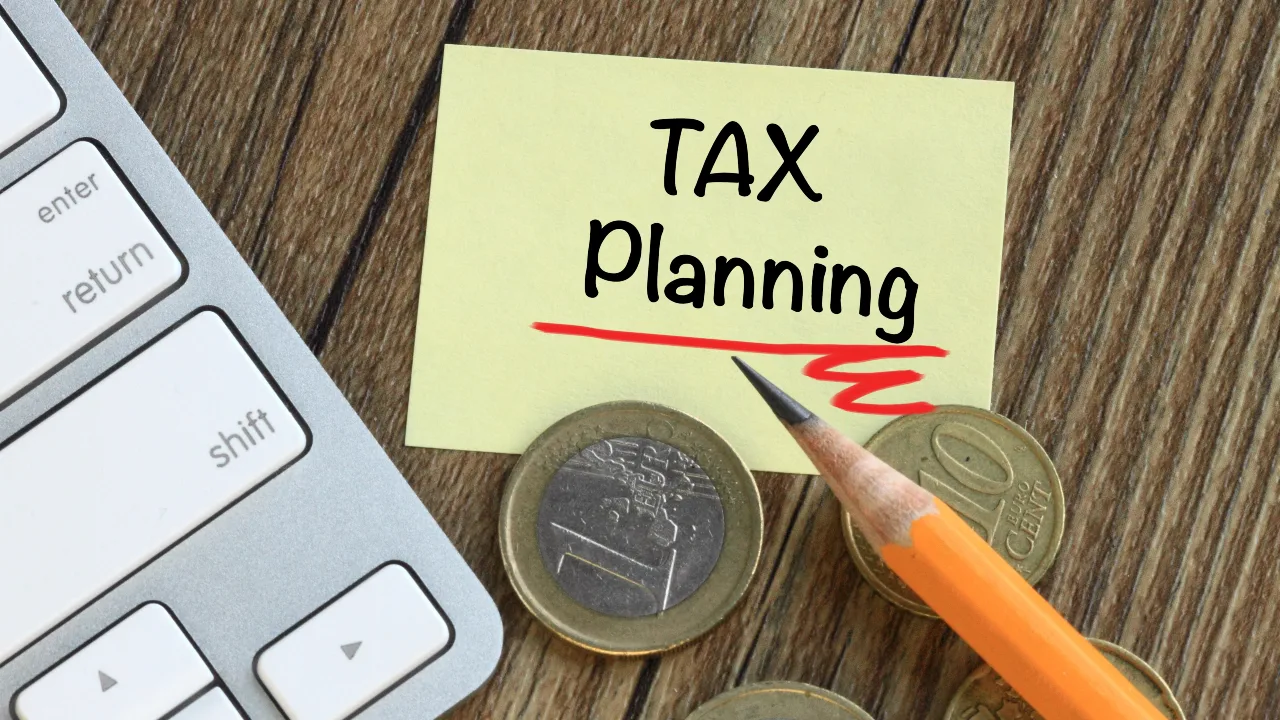 Su una superficie di legno, accanto a una matita, a diverse monete di euro e al bordo della tastiera di un computer, si trova un foglietto adesivo con la scritta "tax planning". Il riferimento va al consolidato fiscale e altre strategie di ottimizzazione finanziaria
