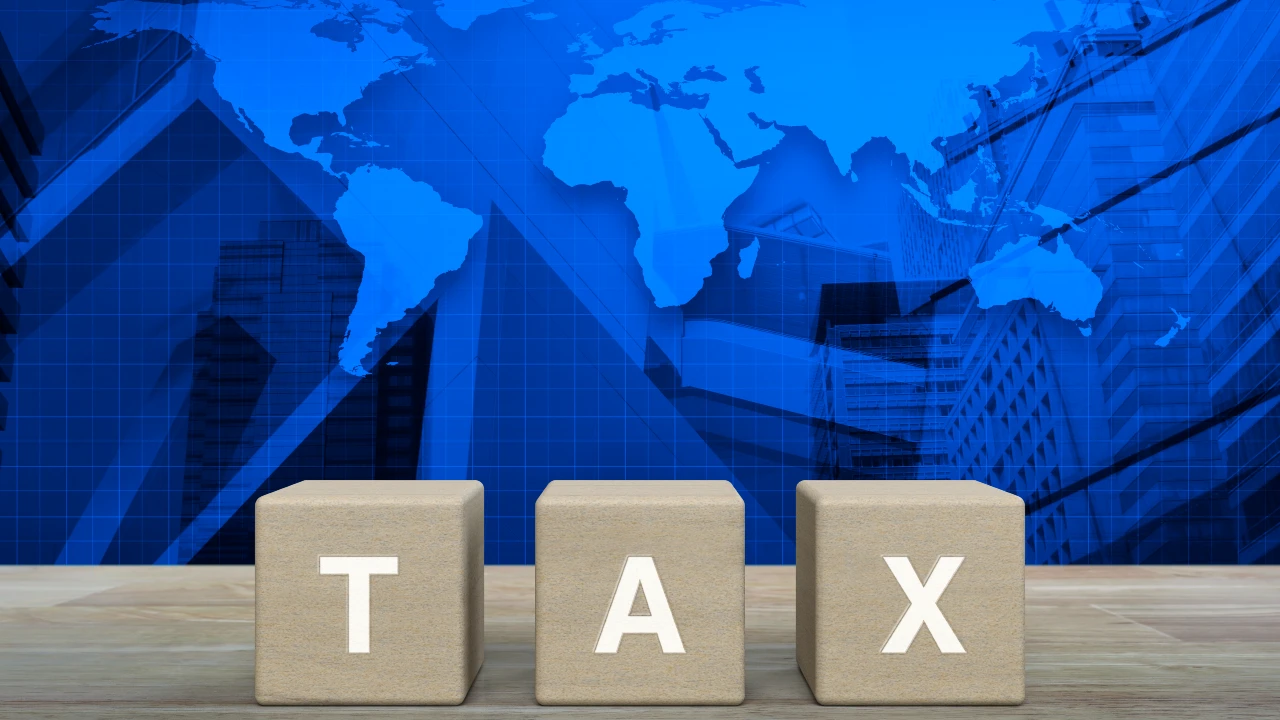 Tre blocchi beige con le lettere "T", "A" e "X" scrivono "Tax" davanti a una mappa del mondo, a simboleggiare le iniziative legate alla global minimum tax.
