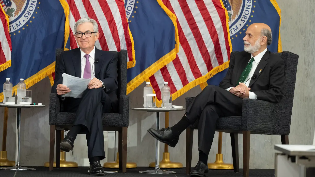 Due uomini in giacca e cravatta siedono sul palco con dei documenti, affiancati da bandiere degli Stati Uniti e dai sigilli della Federal Reserve sullo sfondo. Tra loro c'è un tavolino con bottiglie d'acqua.