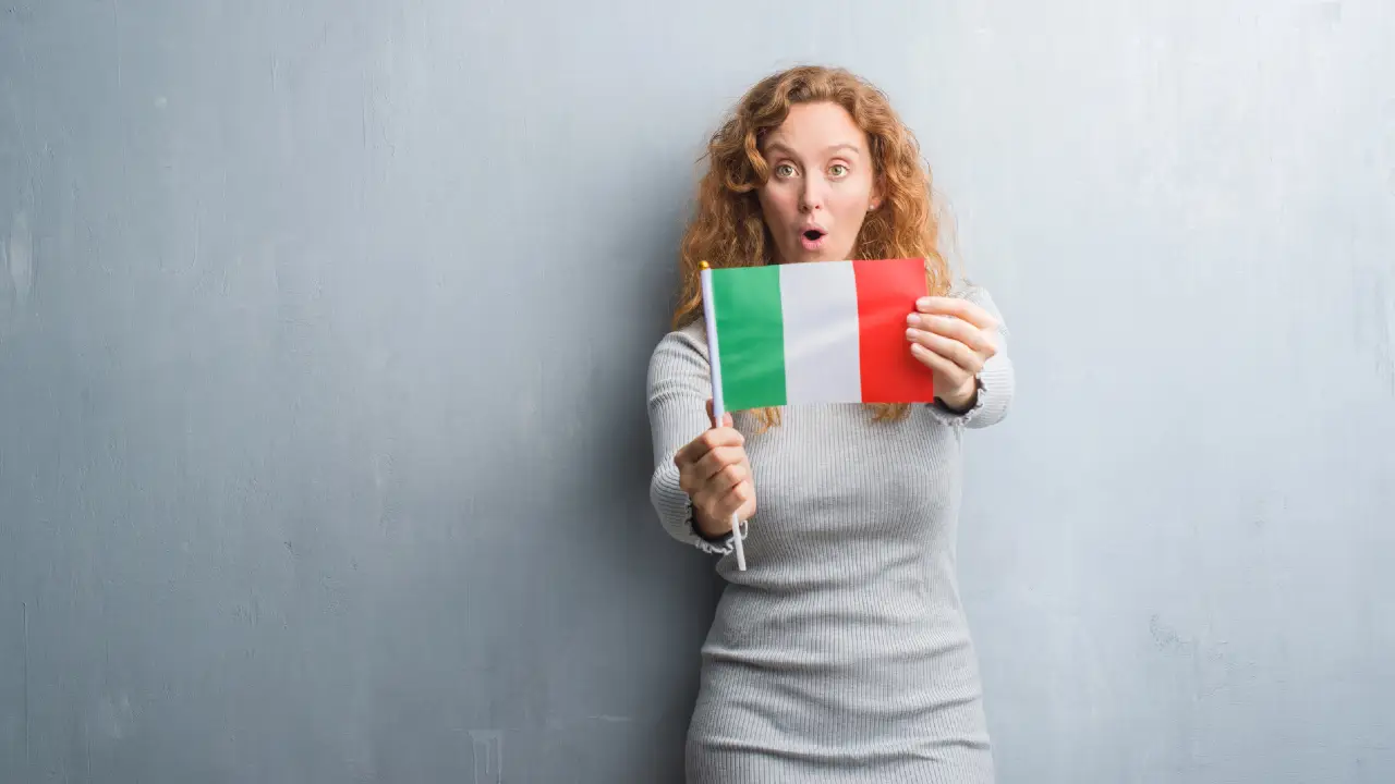Una donna con i capelli ricci che indossa un abito grigio regge una bandiera italiana con un'espressione sorpresa, in piedi di fronte a un semplice muro grigio chiaro.