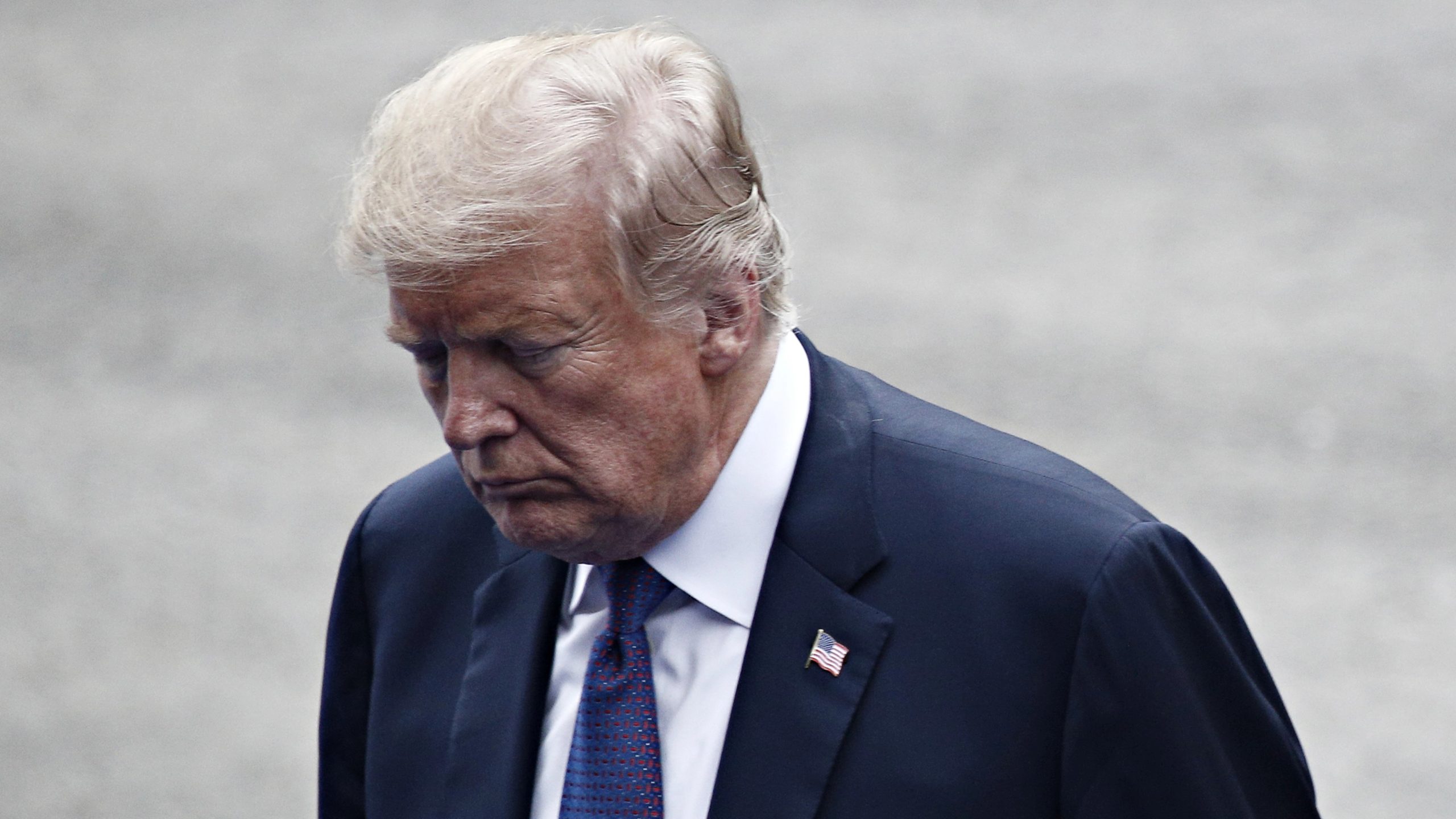Un uomo in abito scuro e cravatta a fantasia guarda verso il basso con espressione seria, all'aperto. Sul bavero della giacca è visibile una spilla con la bandiera americana.