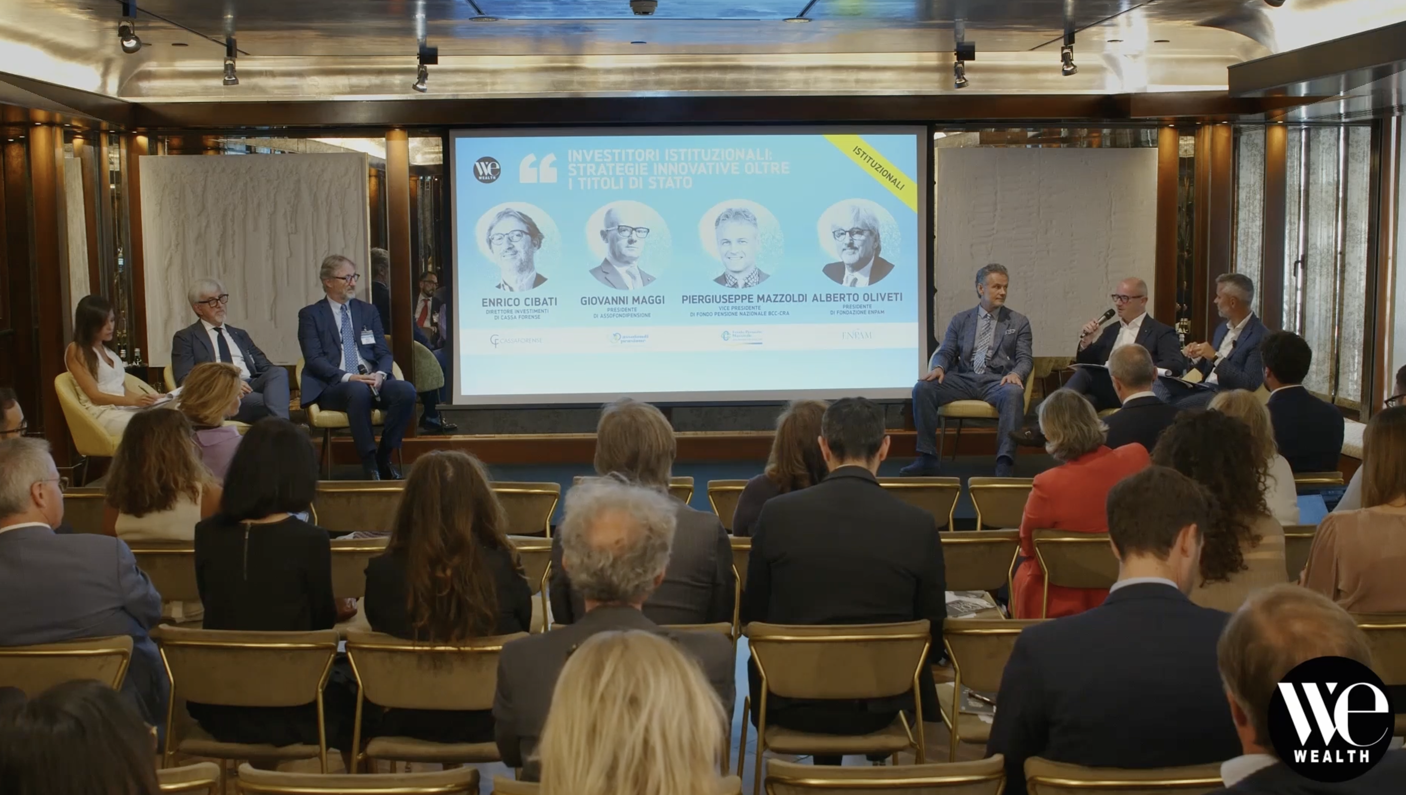 Una tavola rotonda si svolge su un palco davanti a un pubblico. Cinque relatori sono seduti sotto un grande schermo che mostra i loro nomi e le loro foto. Sullo schermo si legge "WEALTH" e si parla di un evento sulle strategie di investimento istituzionali.