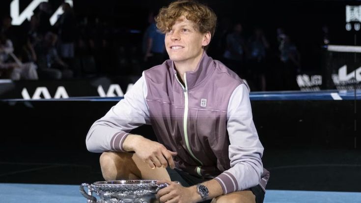 Un giovane con i capelli ricci che indossa una giacca bianca e viola chiaro è inginocchiato su un campo da tennis e sorride mentre posa con un grande trofeo d'argento e mostra con orgoglio il suo orologio. Lo sfondo è poco illuminato con figure sfocate.
