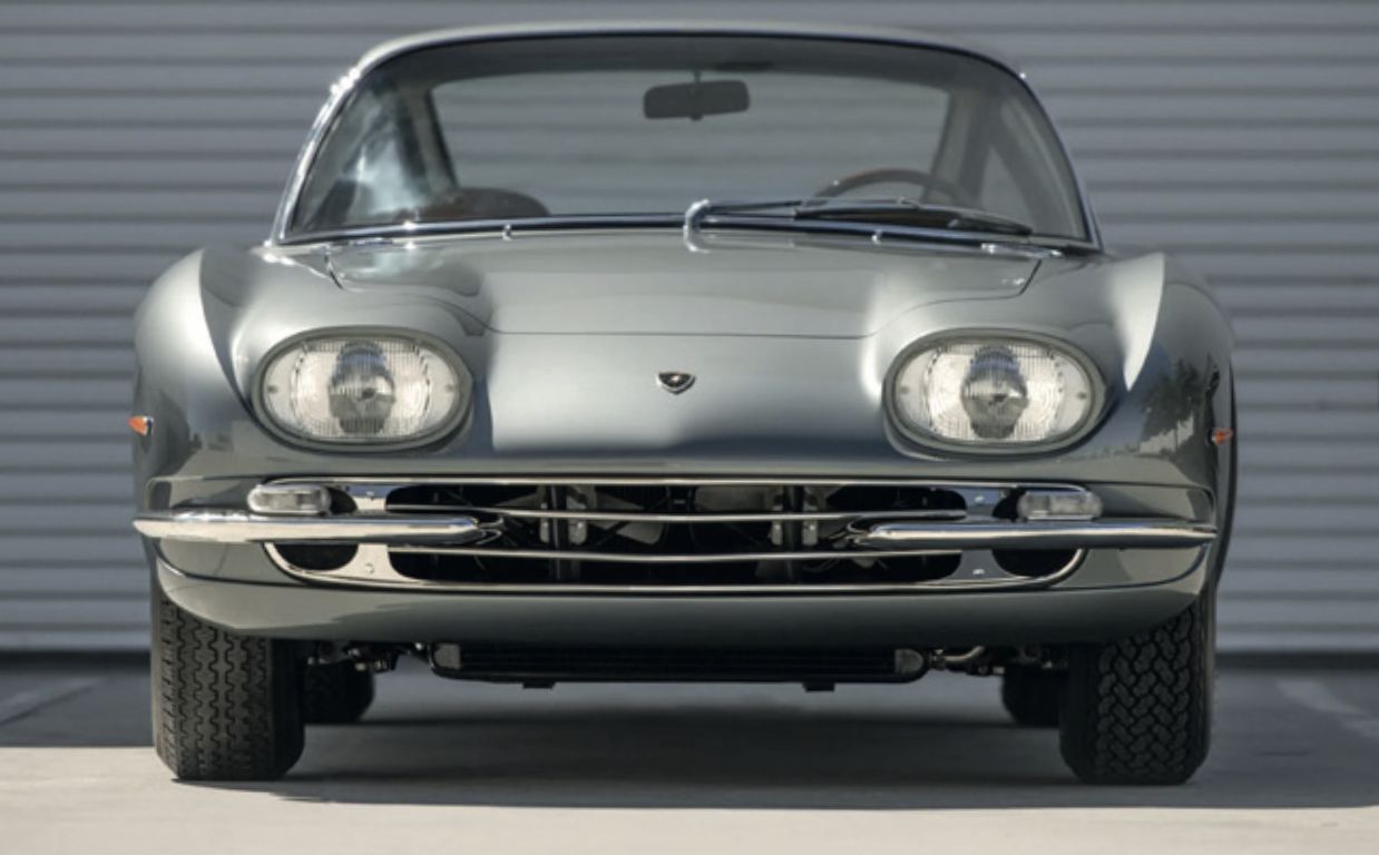 Vista frontale di una classica Lamborghini 400 GT argentata, con fari tondi, cofano basso e un design elegante e sinuoso, parcheggiata su una superficie di cemento di fronte a un muro di metallo ondulato bianco.