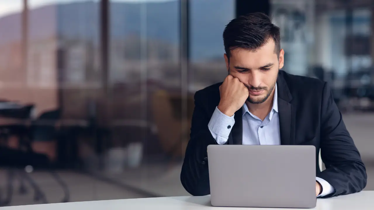 Un uomo in giacca e cravatta siede alla scrivania, appoggiando il mento sulla mano e guardando pensieroso un computer portatile in un ufficio moderno con grandi finestre.