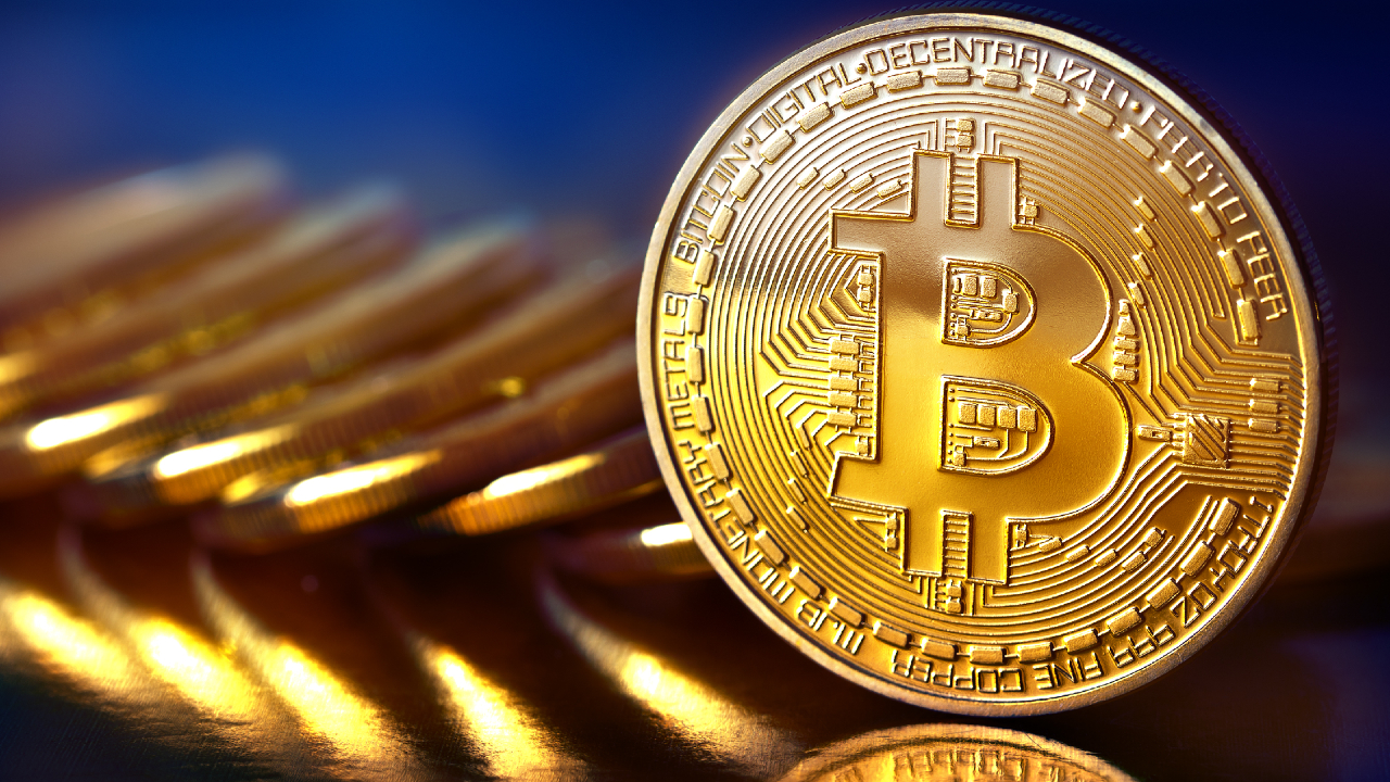 Una moneta Bitcoin d'oro lucida è messa a fuoco, con il simbolo Bitcoin e il disegno del circuito digitale, mentre diverse monete sfocate giacciono sullo sfondo su uno sfondo scuro e colorato.