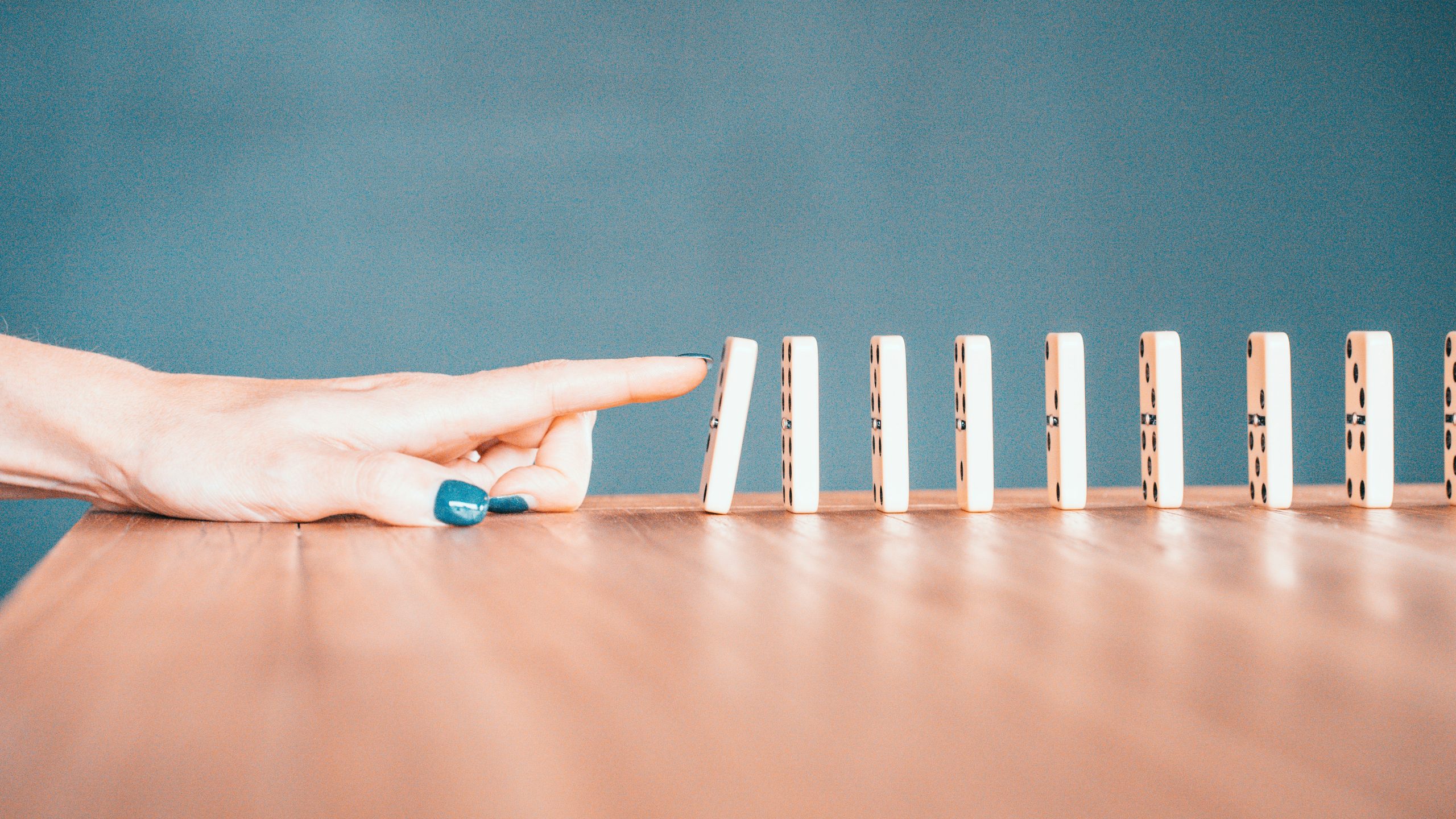 Una mano con smalto blu sta per rovesciare il primo di una fila di domino in piedi su una superficie di legno, suggerendo l'inizio di una reazione a catena.