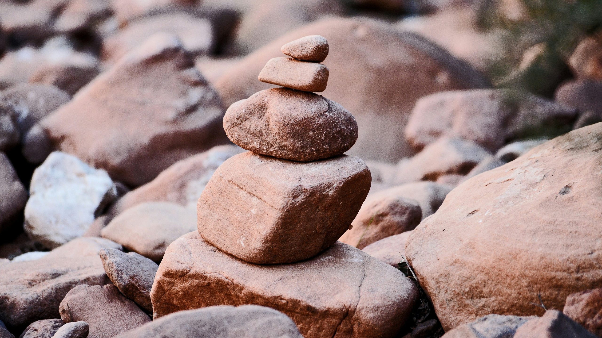 Una piccola pila di cinque rocce equilibrate si trova tra pietre marroni più grandi e sparse all'aperto, creando un semplice cairn di roccia su uno sfondo naturale sfocato.