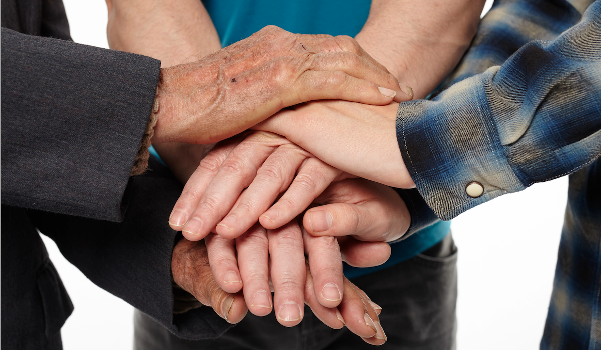 Quattro mani di età e tonalità della pelle diverse impilate l'una sull'altra, a simboleggiare l'unità e il lavoro di squadra, su uno sfondo bianco.