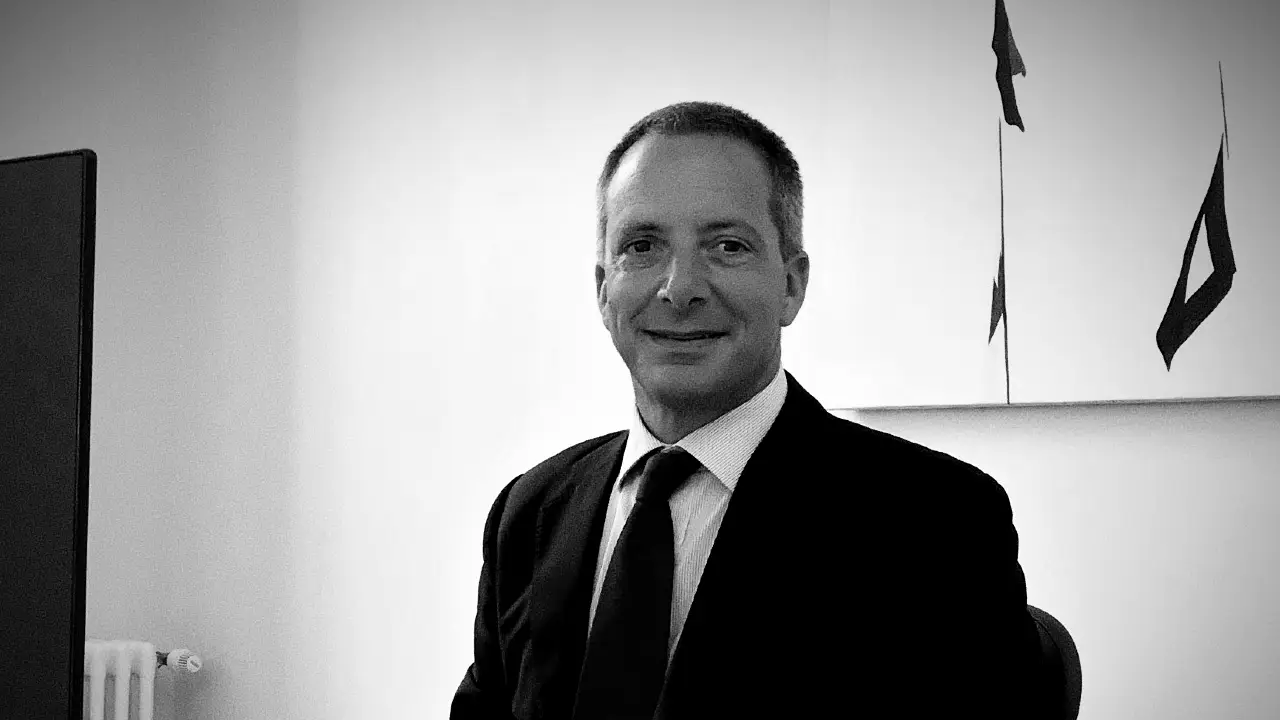 Un uomo in giacca e cravatta siede in un ufficio e sorride alla telecamera. L'immagine è in bianco e nero, con opere d'arte astratte e un radiatore visibili sullo sfondo.