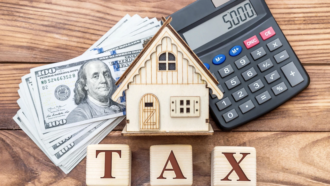 Un modello di casa in legno, una pila di banconote da 100 dollari, una calcolatrice che indica 5000 e blocchi di legno con la scritta "Tax" sono disposti su una superficie di legno, a simboleggiare la tassa di proprietà dell'Ici e dell'Imu.