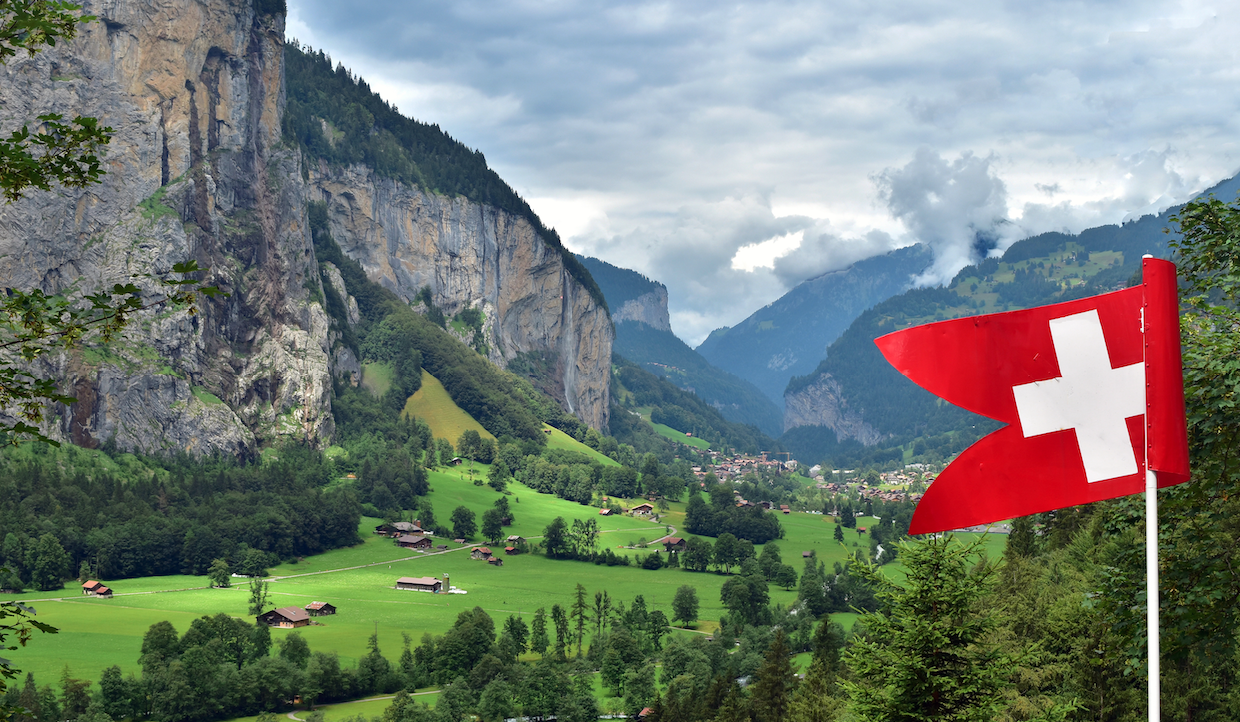 Una bandiera svizzera sventola in primo piano, affacciandosi su una valle verde e rigogliosa con case sparse, ripide scogliere e montagne boscose sotto un cielo nuvoloso in Svizzera.