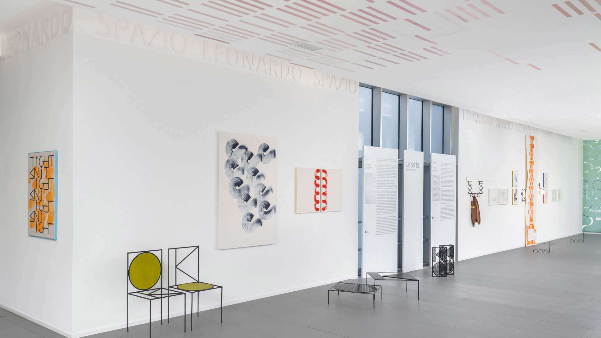 Una galleria d'arte moderna con dipinti astratti, sedie geometriche e alte vetrate. Opere d'arte e pannelli informativi sono allineati su una parete bianca, con Leonardo Assicurazioni arte e disegni colorati sul soffitto e sul pavimento.