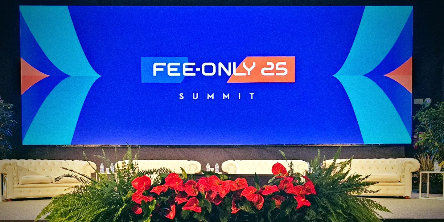 Un grande palcoscenico con divani bianchi, piante e fiori rossi davanti a uno schermo blu che mostra a caratteri cubitali "FEE-ONLY 25 SUMMIT".