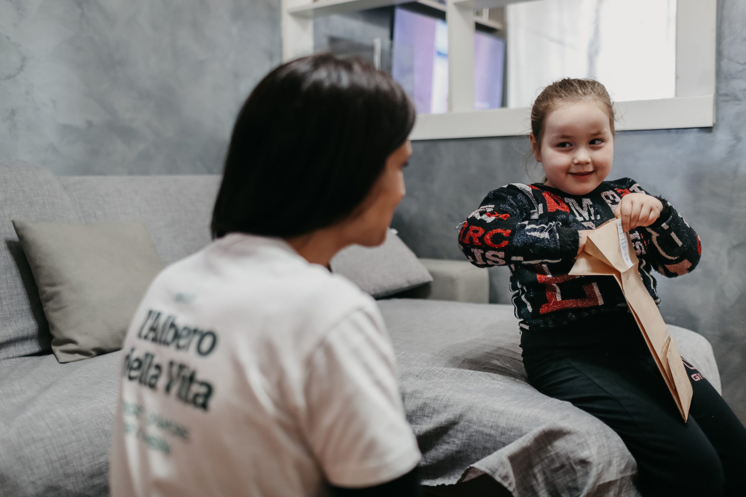 Una bambina seduta su un divano apre una busta marrone e sorride a un adulto vicino, che indossa una maglietta bianca con un testo, forse legato a La rondine fonazione l'albero della vita.