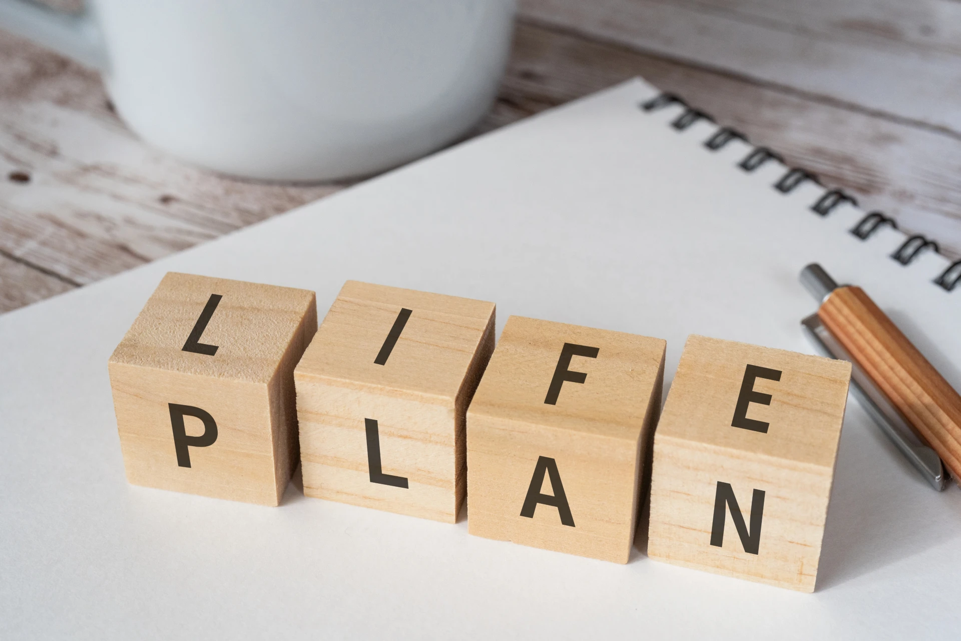 Blocchi di legno scrivono "LIFE PLAN" su una superficie bianca accanto a un quaderno a spirale e a una penna, evocando i temi dell'organizzazione e dello sviluppo personale, perfetti per esplorare assicurazioni e pianificazione finanziaria di nuova generazione.