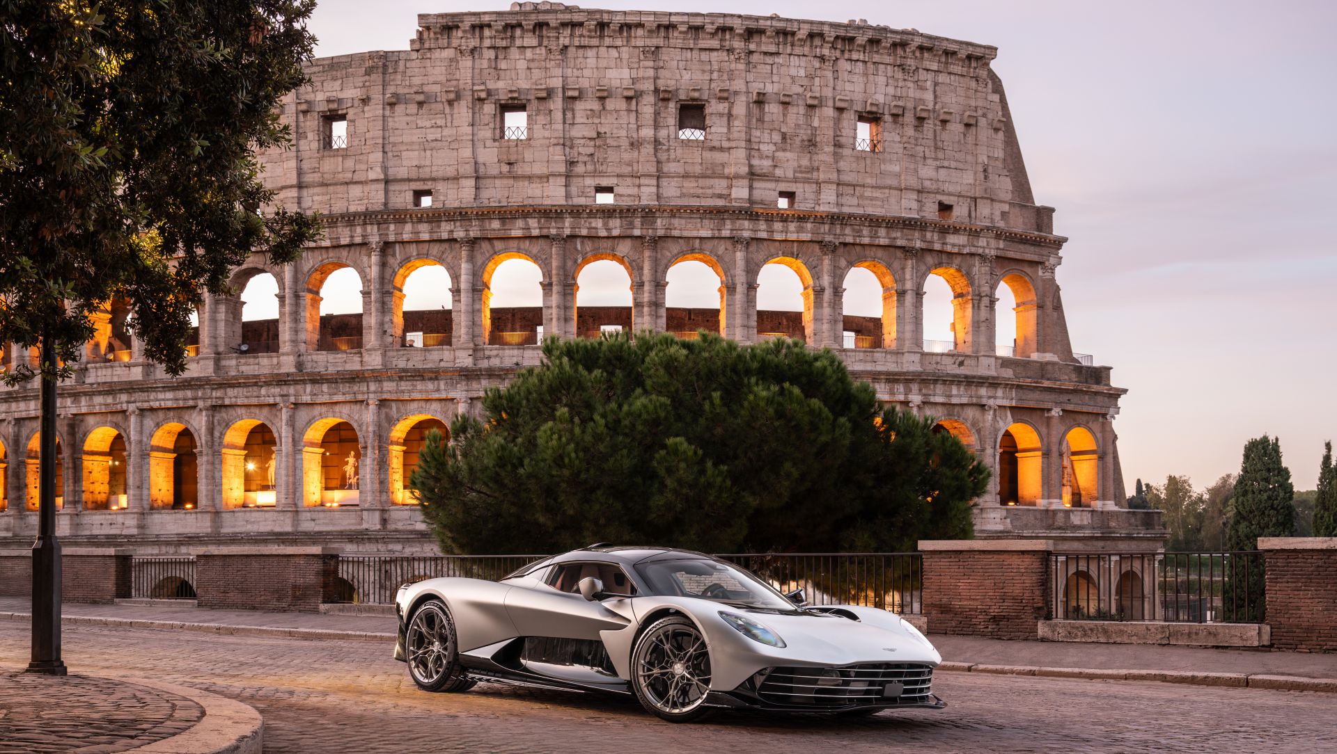 Un'auto sportiva argentata è parcheggiata su una strada di ciottoli di fronte al Colosseo illuminato a Roma al tramonto, con gli alberi che oscurano parzialmente parte dell'antica struttura.