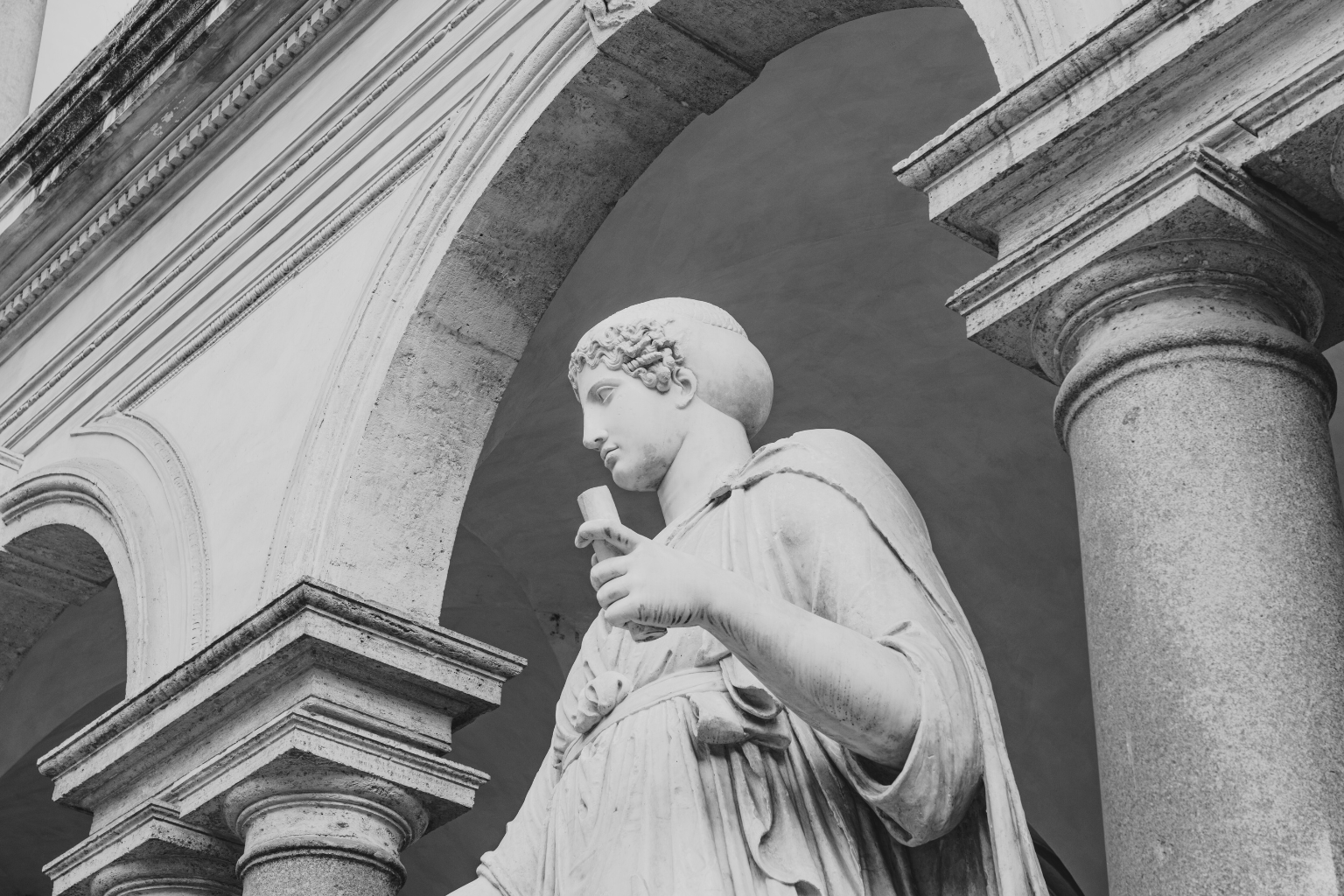 Foto in bianco e nero di una statua di marmo in abiti romani, sotto grandi archi e colonne di pietra, che guarda di lato tenendo in mano un oggetto: un'immagine che ricorda la visione senza tempo di Blue Ocean Family Office.