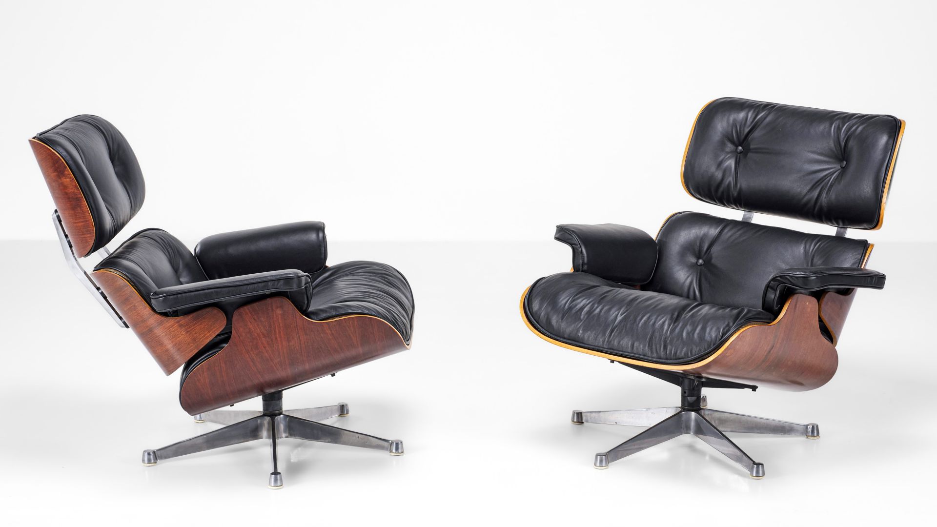 Due iconiche poltrone da salotto della metà del secolo scorso, con rivestimenti in pelle nera, strutture in legno curvato e basi girevoli in metallo - pezzi perfetti per elevare qualsiasi design asta settembre - sono posizionate una di fronte all'altra su uno sfondo bianco.