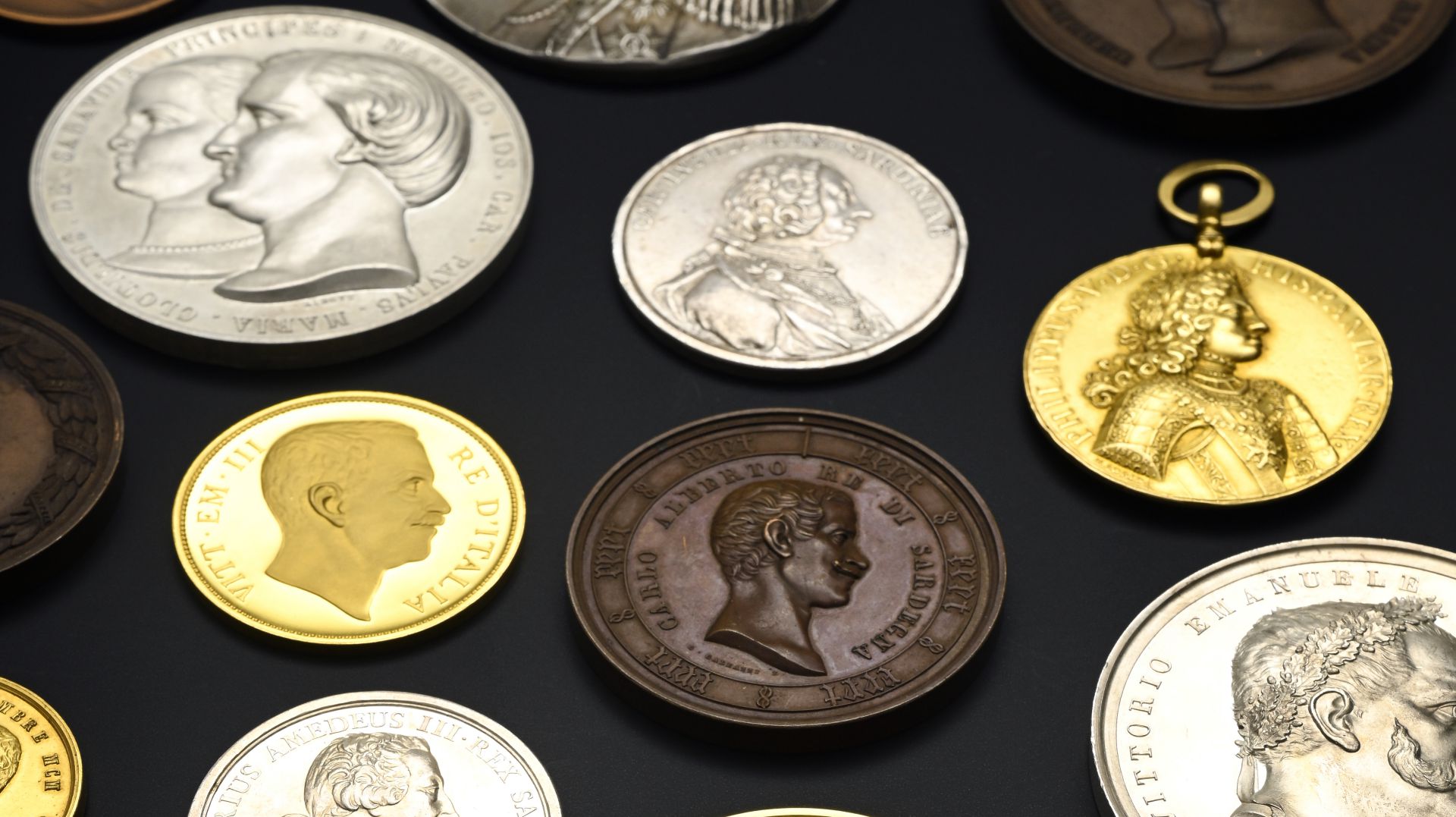 Una varietà di monete d'epoca e medaglie Savoia con diversi ritratti e iscrizioni sono disposte su una superficie scura, con colori argento, oro e bronzo.