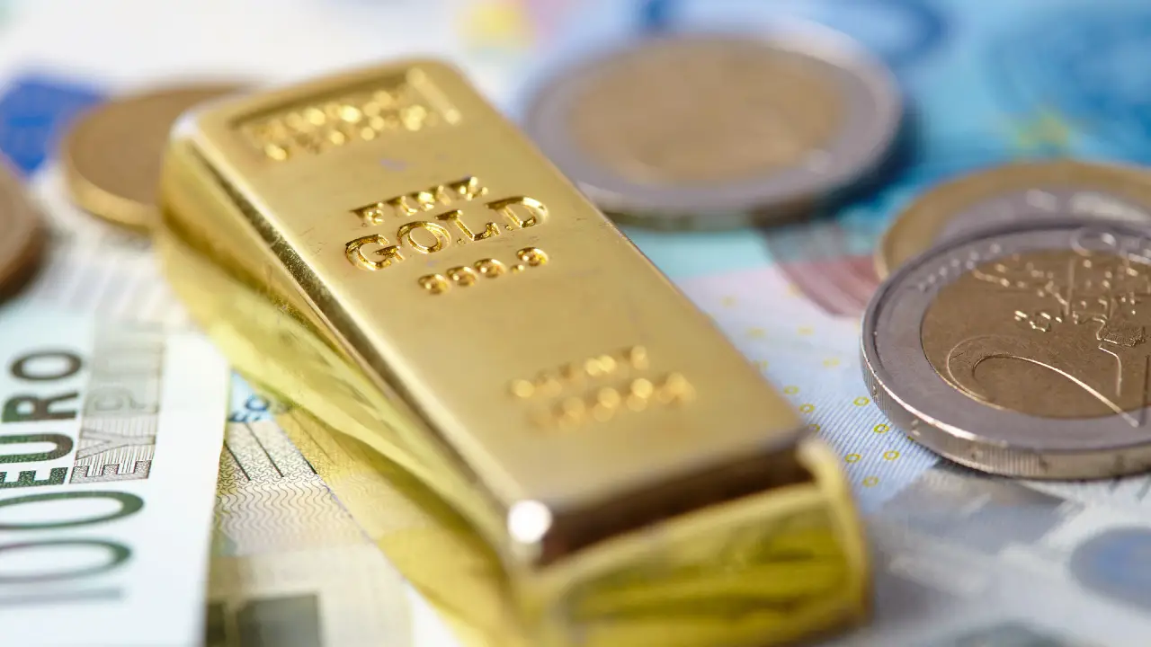 Un lingotto d'oro con l'etichetta "ORO FINE 999,9" poggia su banconote in euro, circondato da diverse monete in euro.