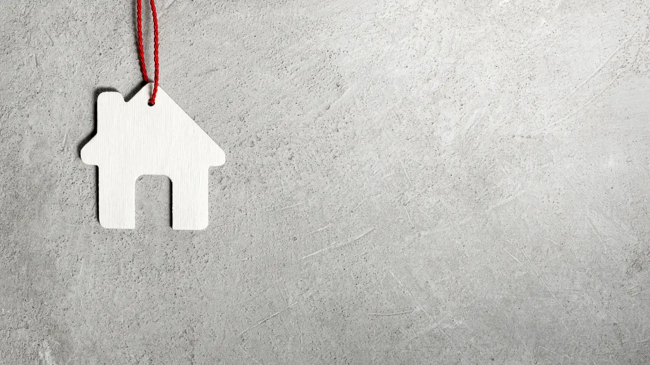 Un piccolo ornamento bianco a forma di casa in legno è appeso a un filo rosso su uno sfondo grigio con un ampio spazio vuoto a destra.